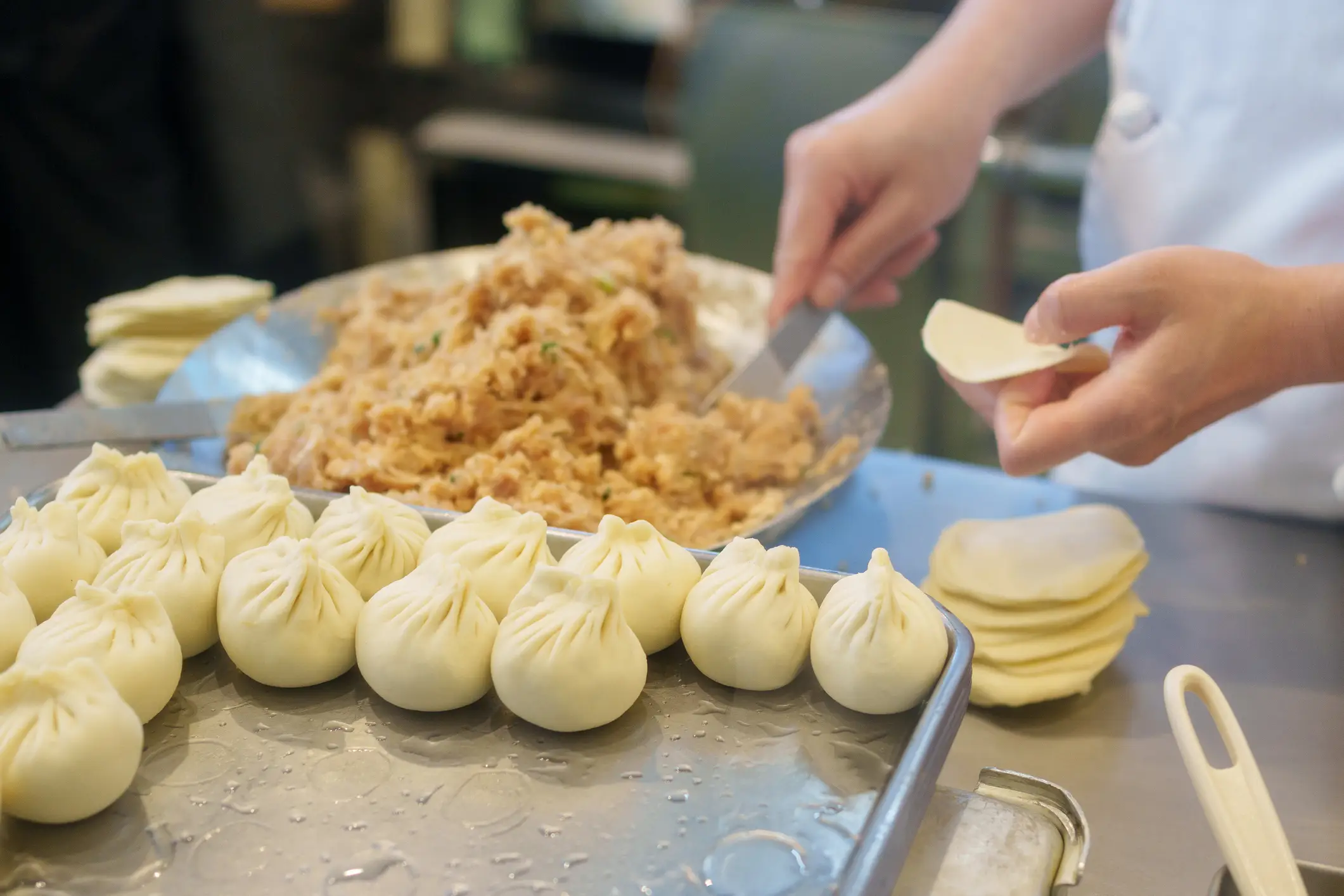 Zubereitung von Dim Sum: Gefüllte Teigtaschen auf Tablett, Hände bereiten Füllung vor, Teigscheiben liegen bereit.