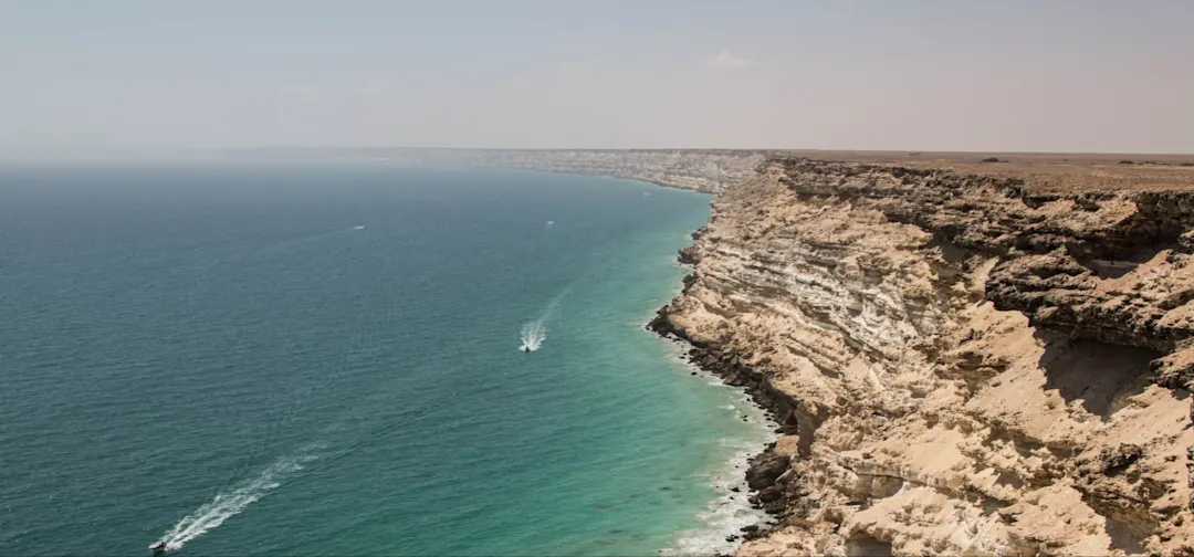 Eine schroffe, felsige Klippe erstreckt sich entlang der Küste und trifft auf klares, türkisfarbenes Wasser unter einem dunstigen Himmel. Mehrere kleine Boote hinterlassen weiße Spuren auf dem ruhigen Meer in der Nähe des Fußes der Klippen.