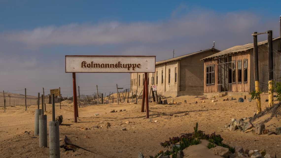 Kolmannskuppe, deutsche Schreibweise von Kolmanskop, Schild mit verlassenen Gebäuden in der Geisterstadt eines ehemaligen Diamantenabbaugebiets in der Namib-Wüste von Namibia.