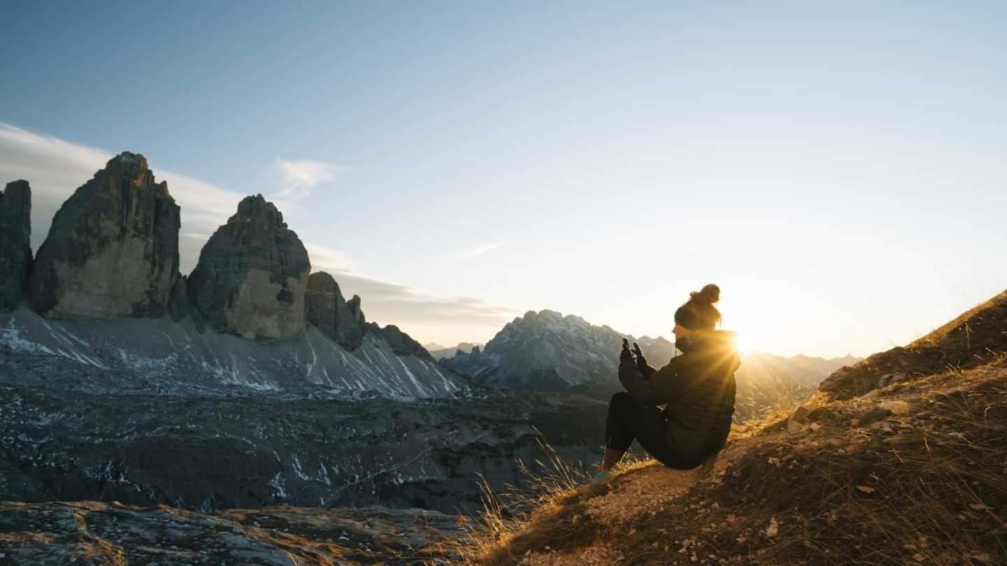Randonneuse assise sur un flanc de montagne au levée du soleil en train de prendre en photo le paysage.