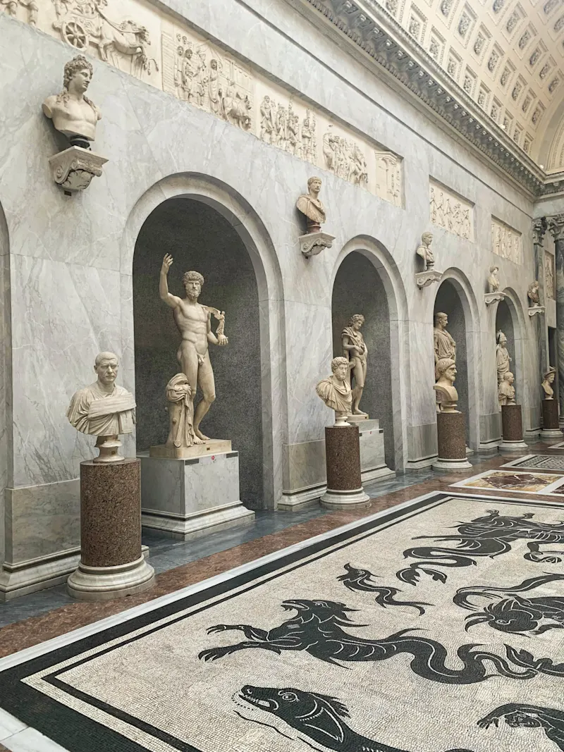 Galerie de musée avec statues antiques romaines dans des alcôves en marbre et mosaïque décorative au sol.