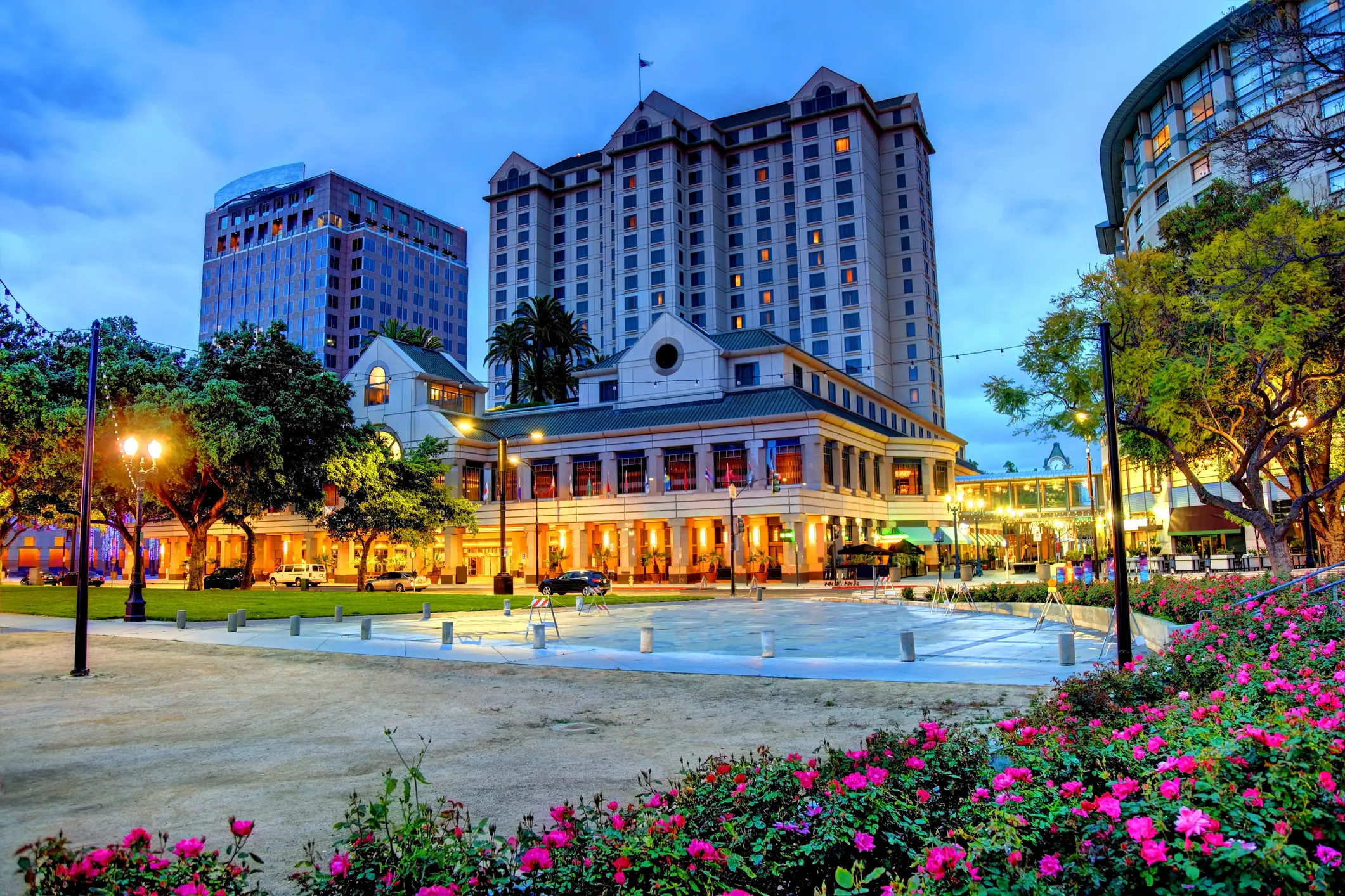 Eine Stadtlandschaft in der Abenddämmerung mit einem hohen Hotelgebäude, einem historischen Gebäude mit warmen Lichtern, einem Park mit blühenden Blumen und Bäumen im Vordergrund und einem bewölkten blauen Himmel darüber.