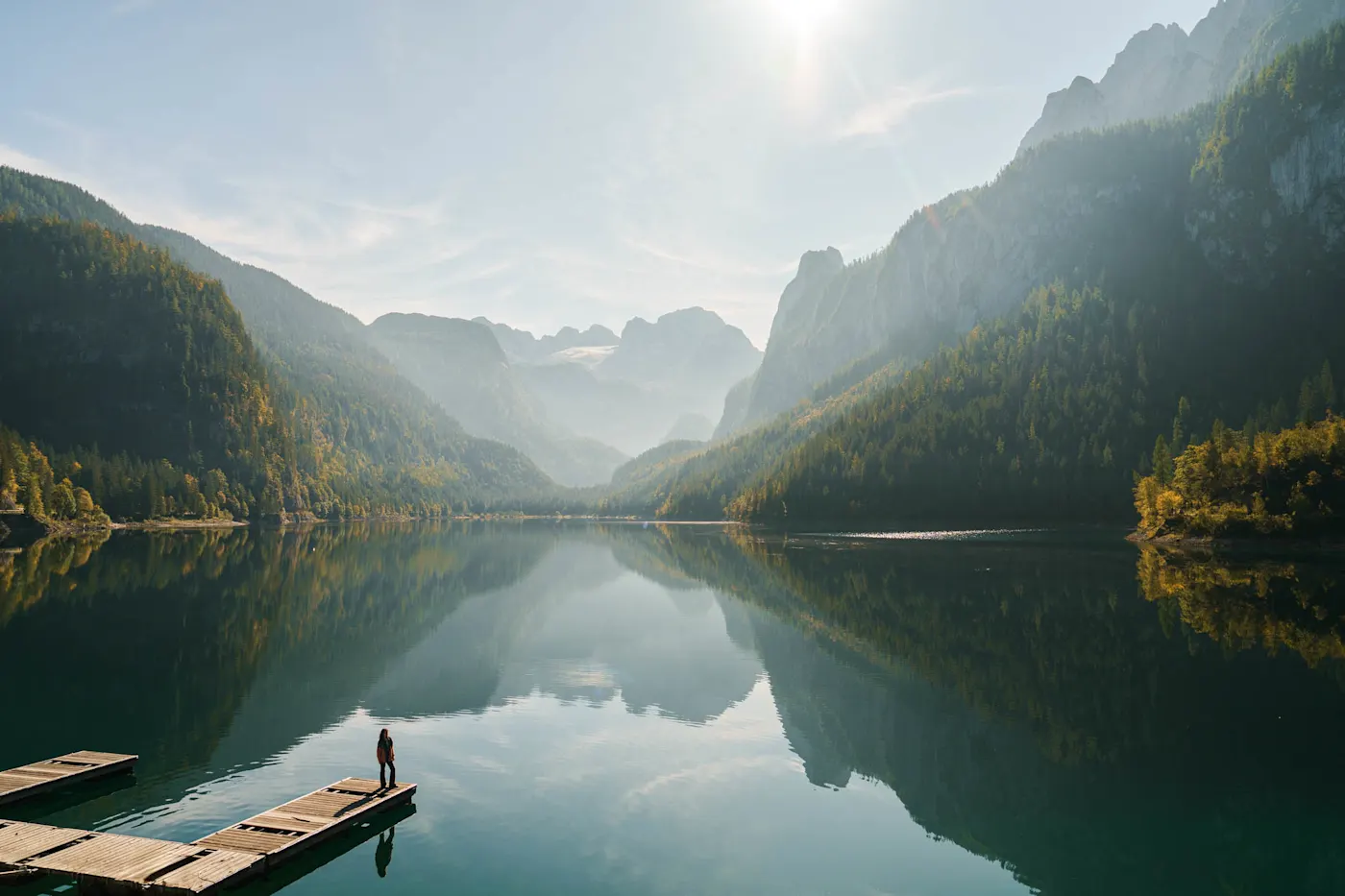 Person standing on wooden dock overlooking serene mountain lake with perfect reflections in morning light.