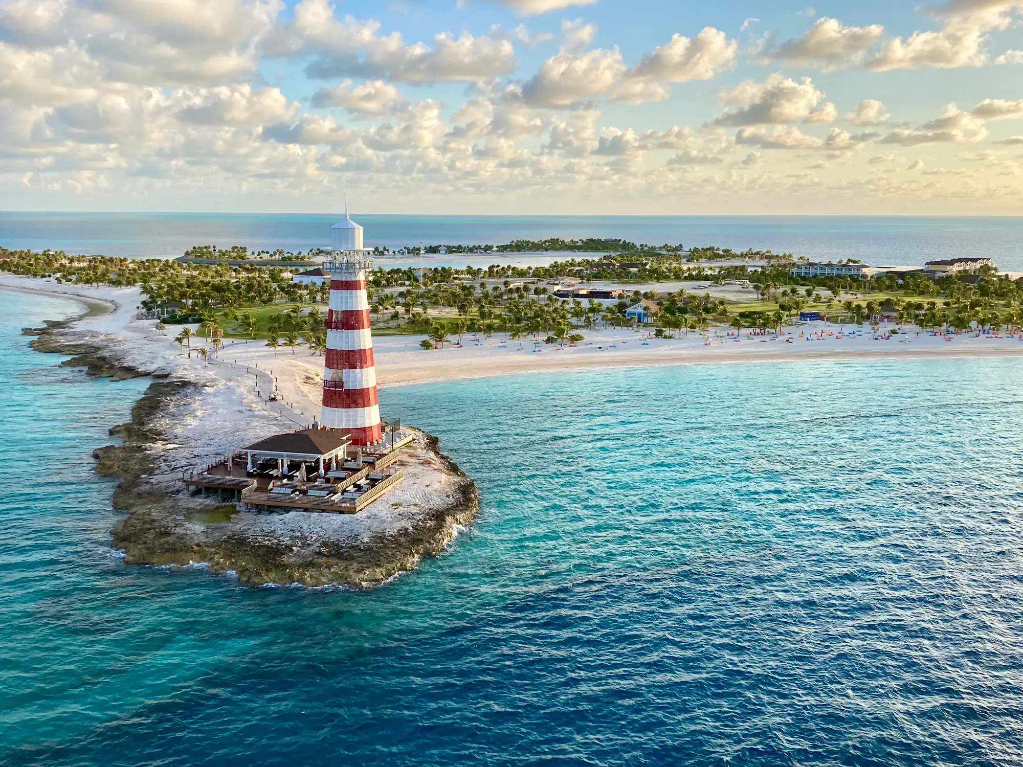 Rot-weiß gestreifter Leuchtturm auf felsigem Vorsprung im türkisblauen Meer, mit Palmenstrand und Inselresort im Hintergrund.
