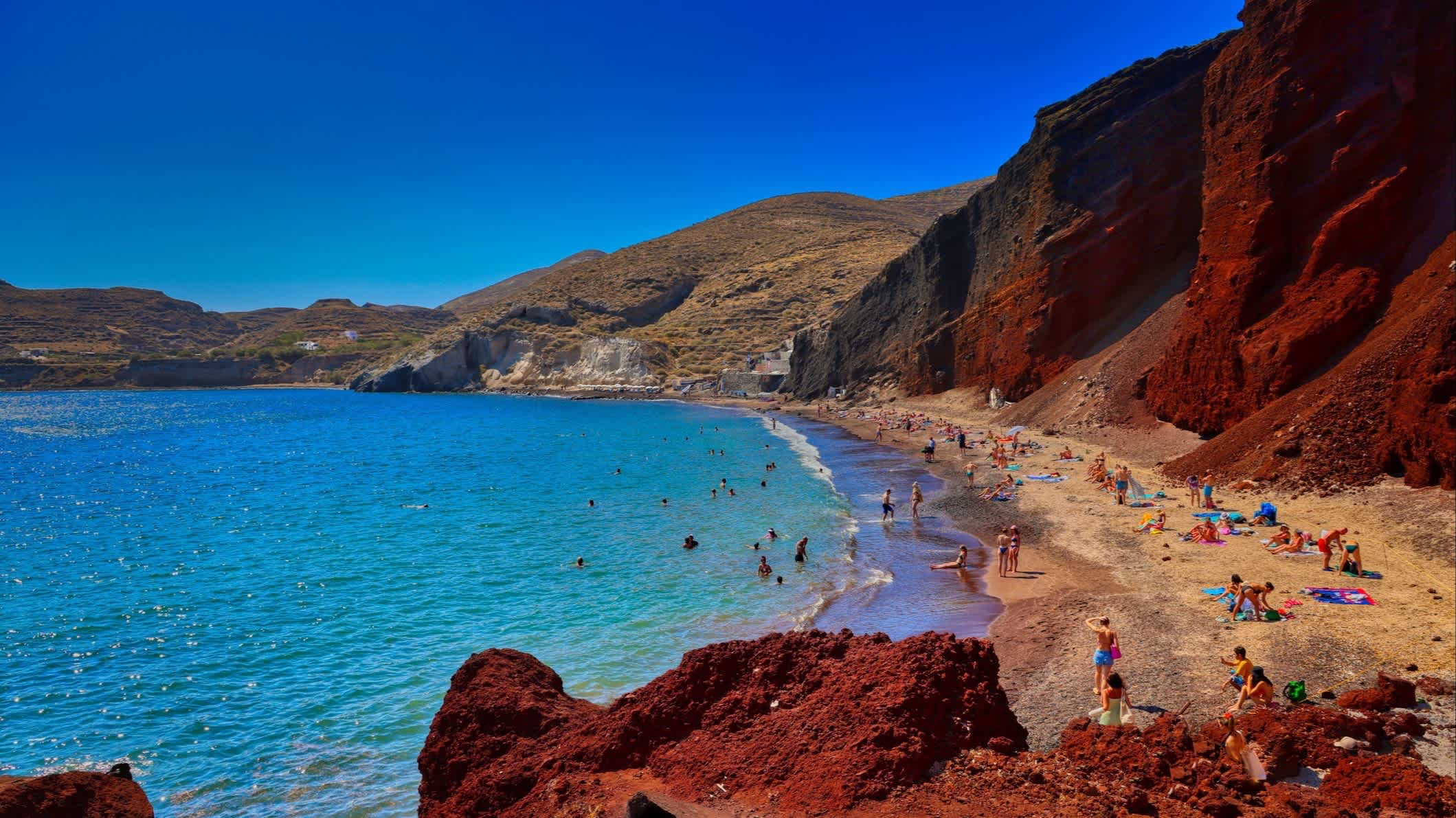 Blick zum Red Beach auf Santorini, Griechenland mit imposanten roten Klippen sowie Menschen am Strand.