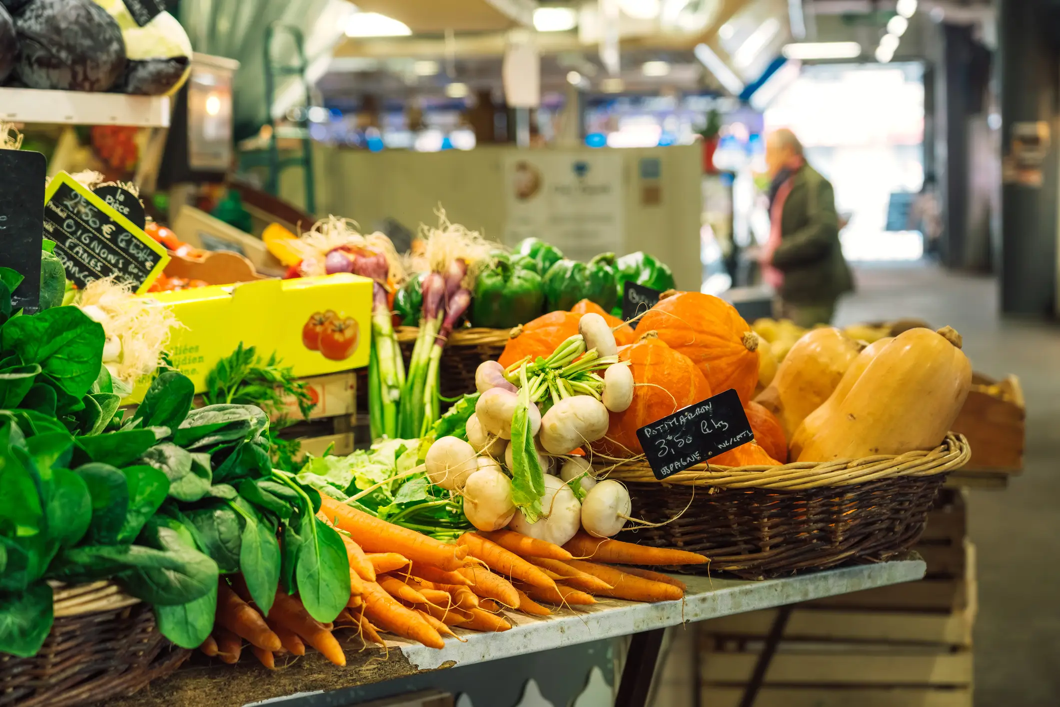 Fresh produce display at indoor market with carrots, radishes, pumpkins and leafy greens in wicker baskets.