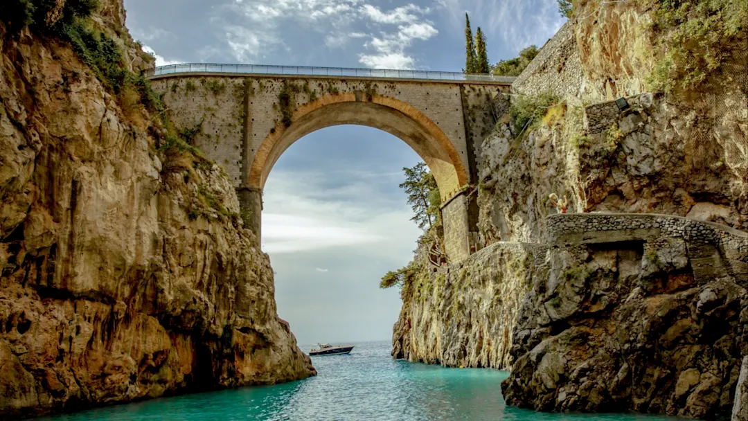 Blick auf eine alte Steinbrücke in einer kleinen Bucht zwischen großen Felsen und darunter das Meer
