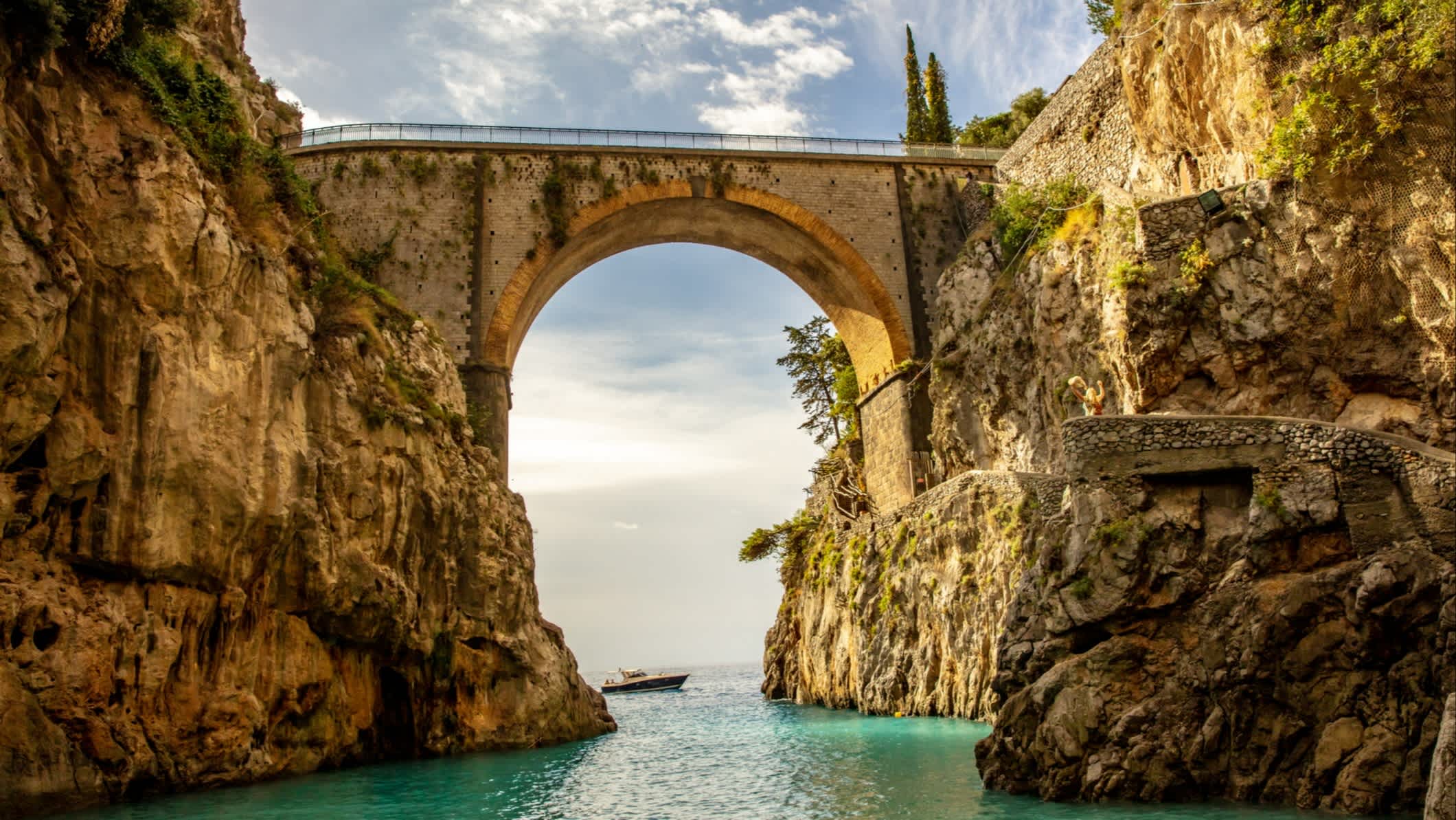 Blick auf eine alte Steinbrücke in einer kleinen Bucht zwischen großen Felsen und darunter das Meer