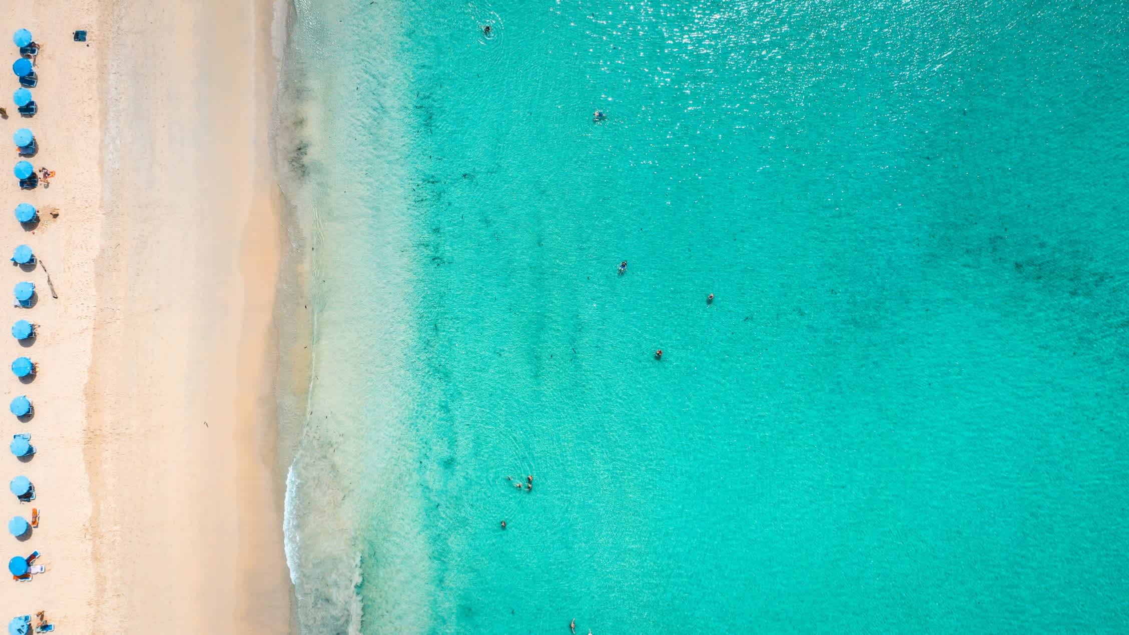 Luftaufnahme des Kata Noi Beach auf Phuket mit türkisblauem Wasser, Sandstrand und blauen Sonnenschirmen.