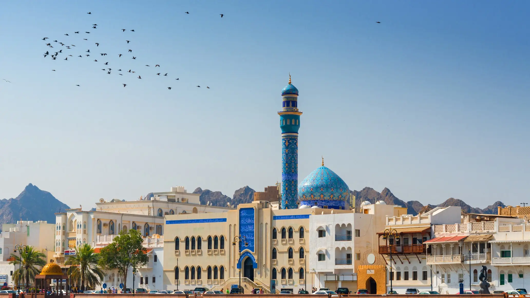 Mosquée avec minaret bleu décoré et dôme turquoise sous un ciel clair, oiseaux en vol, montagnes en arrière-plan, Oman.