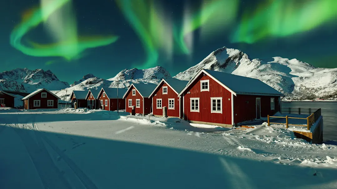 Nordlichter über roten Holzhäusern im Schnee. Reine, Lofoten, Norwegen