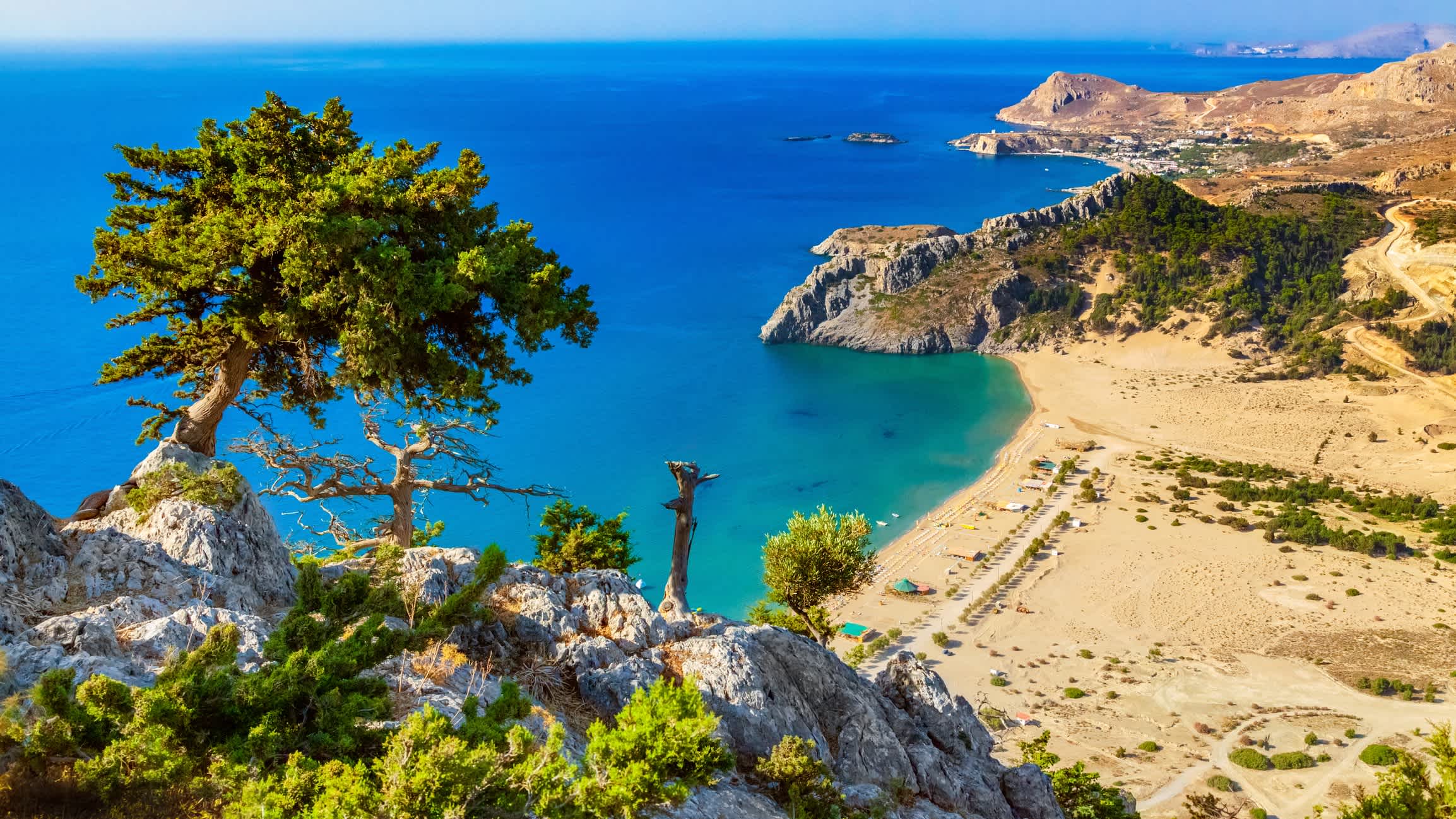 Ein Panorama des Strandes von Tsampika in Rhodos, Griechenland mit Blick auf den goldenen Sand und das blaugrüne Meer sowie Bergen im Hintergrund und Bäume.