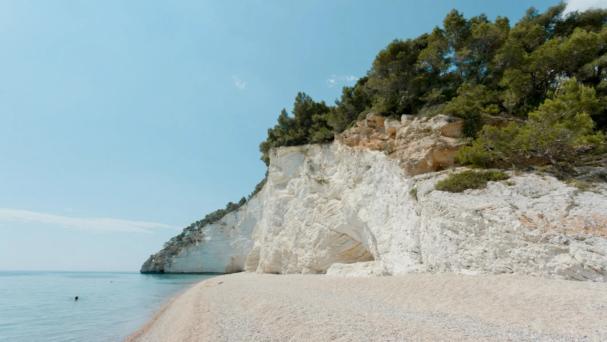 Weiße Kalksteinklippen mit grüner Vegetation an einem Kiesstrand mit türkisblauem Meerwasser unter klarem Himmel.