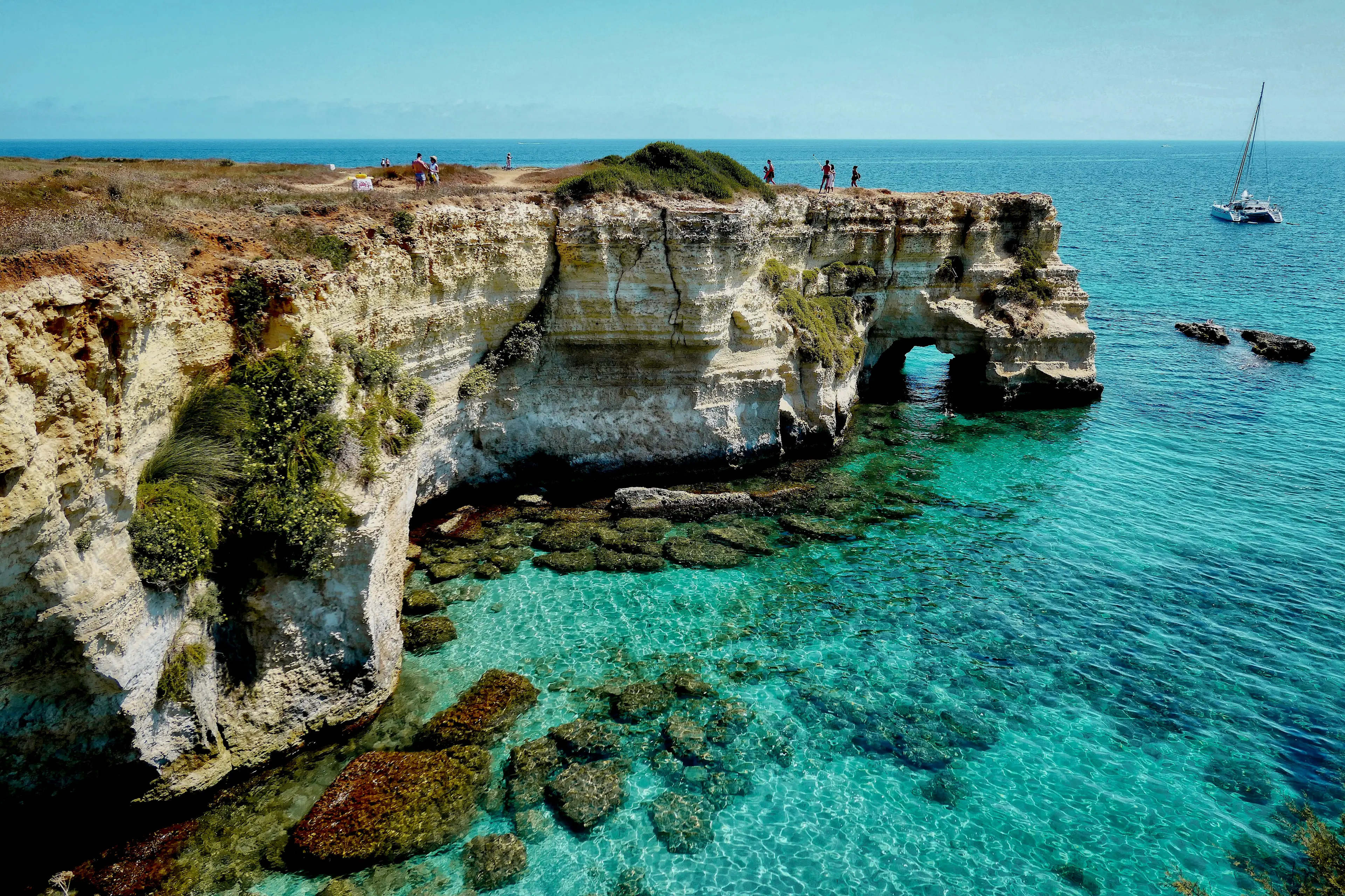 Klares türkisfarbenes Wasser umgibt zerklüftete Kalksteinfelsen mit grünem Gebüsch, Menschen, die am Ufer entlang spazieren, und ein Segelboot, das in der Ferne unter einem strahlend blauen Himmel treibt.