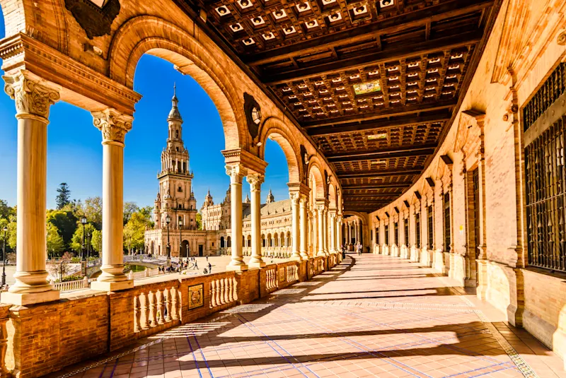 Gewölbter Gang mit kunstvollen Kacheln und Holzdecke auf der Plaza de España in Sevilla, Spanien, mit einem zentralen Turm und blauem Himmel, der durch die Bögen sichtbar ist. Das Sonnenlicht wirft lange Schatten auf den gefliesten Boden.