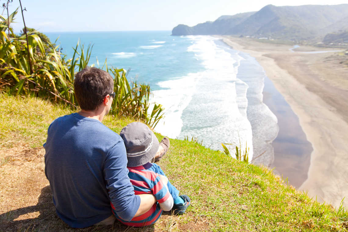 Un père et son enfant photographiés de dos au sommet du rocher de Lion Rock sur la plage de Piha, en Nouvelle-Zélande.