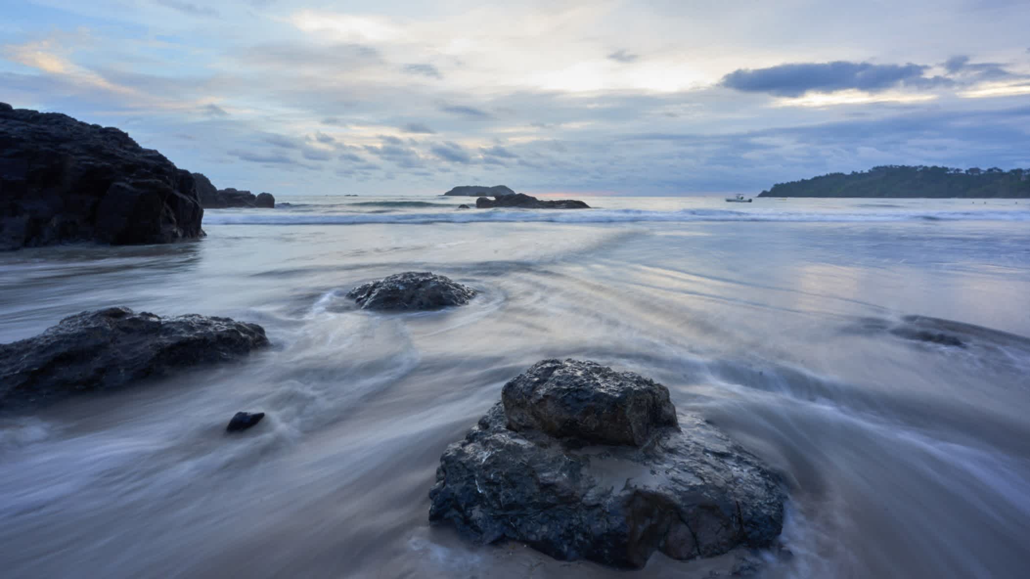 Blick auf den Sonnenuntergang am Strand La Playitas in Quepos, Costa Rica bei Wolken am Himmel und mit schwarzen Felsen. 