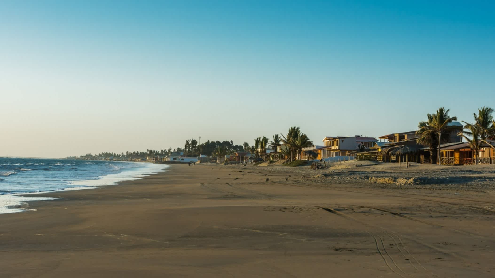 Piura Strand in Colan, Peru mit Blick auf den dunklen Sand des Strandes, bei blauem Himmel und mit Villen und Palmen am Rand.