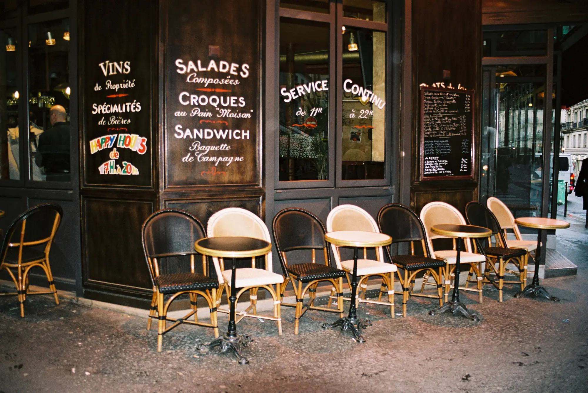 Traditional Parisian café storefront with wicker chairs and tables outside, menu written on windows advertising sandwiches and wine.