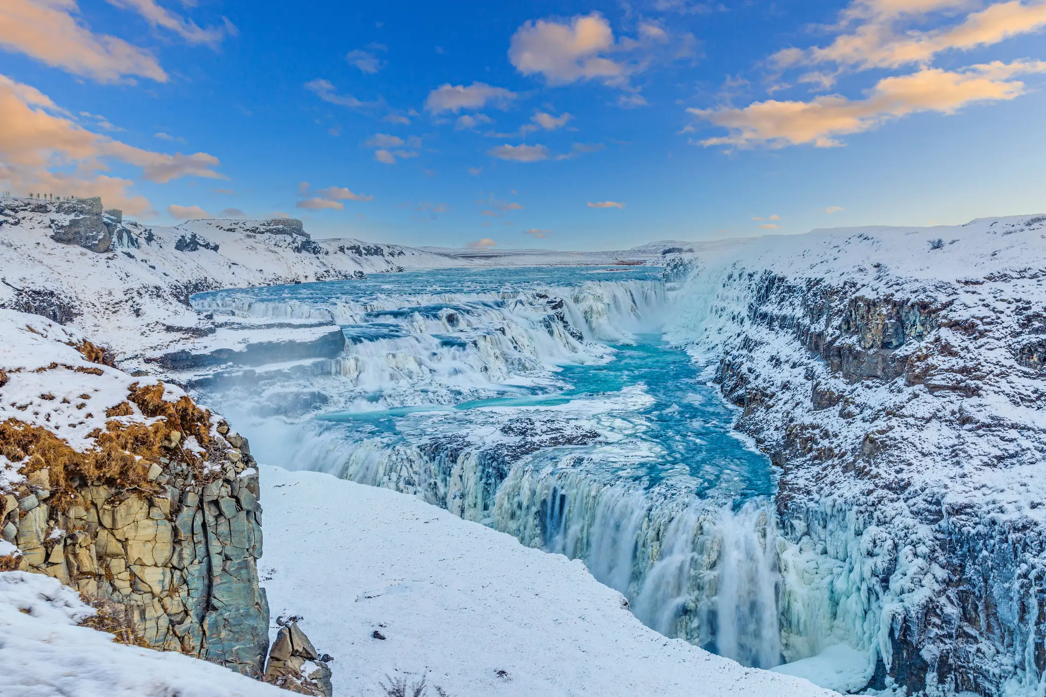 Cascade Gullfoss en Islande en hiver, avec eaux turquoise contrastant avec paysage enneigé sous ciel bleu nuageux.