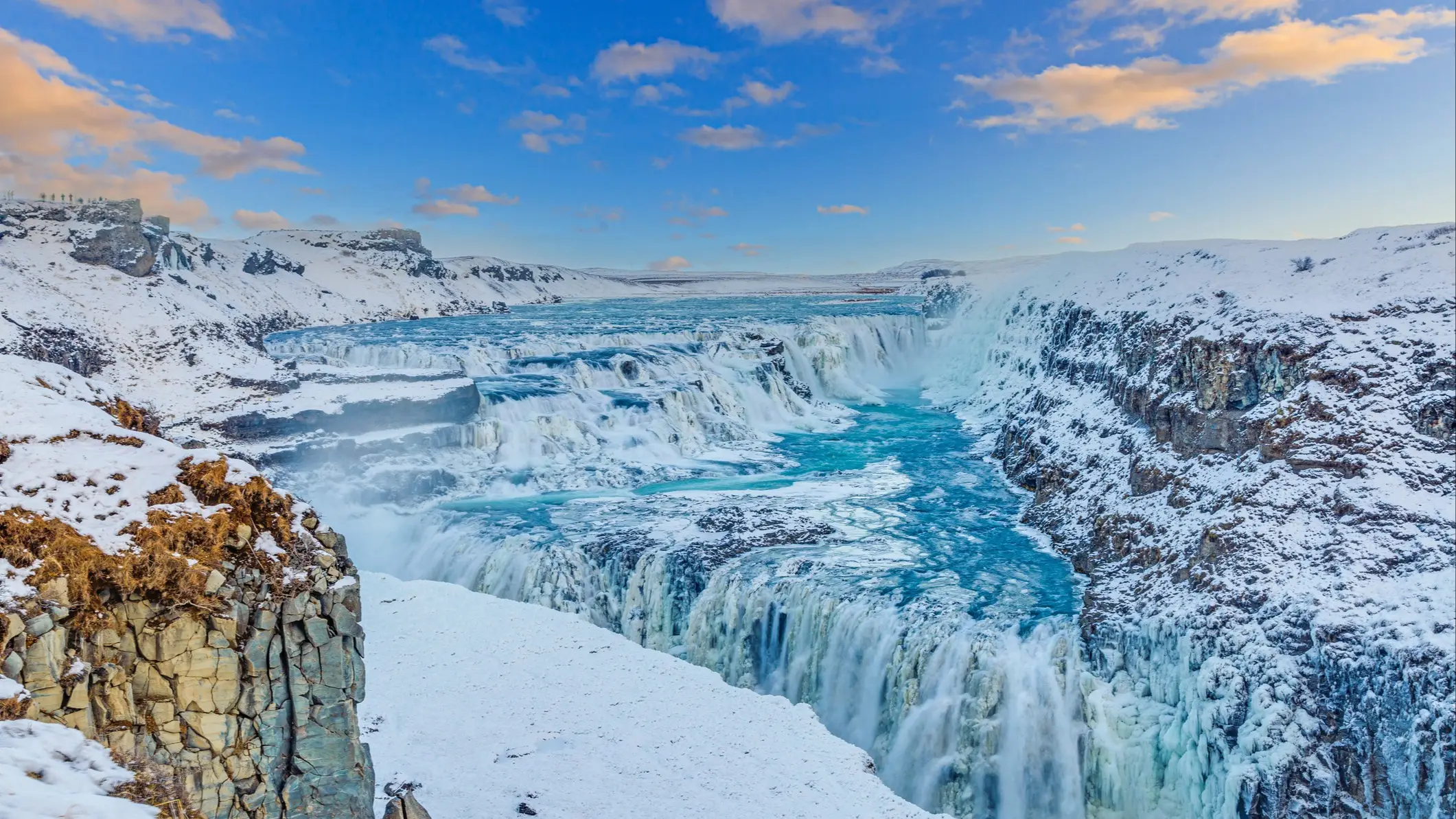 Cascade Gullfoss en Islande en hiver, avec eaux turquoise contrastant avec paysage enneigé sous ciel bleu nuageux.
