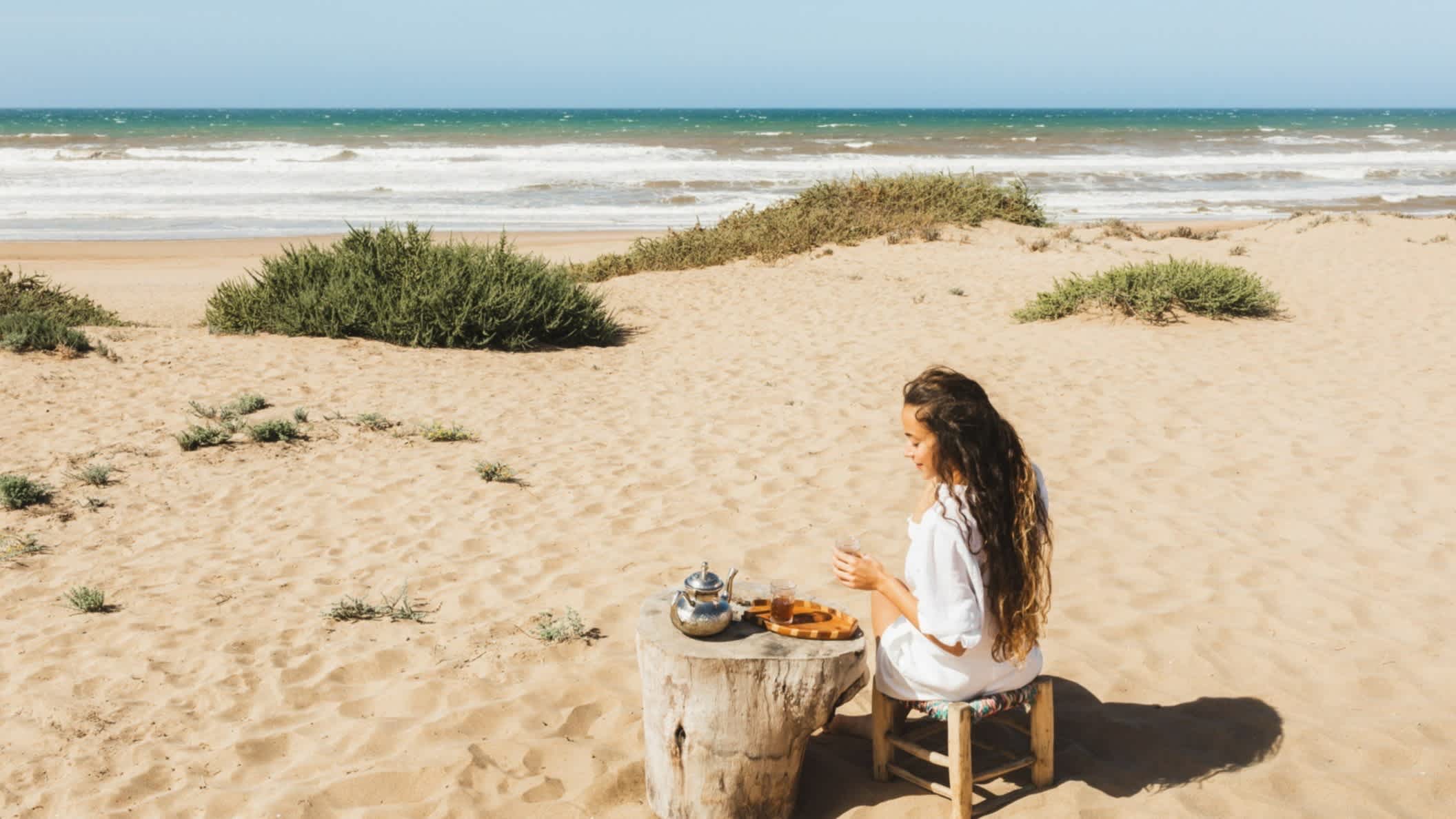 Frau genießt marokkanischen Tee am Sandstrand des Kaf Lahmam, Tanger-Tétouan-Al Hoceïma, in Marokko bei sonnigem Wetter.

