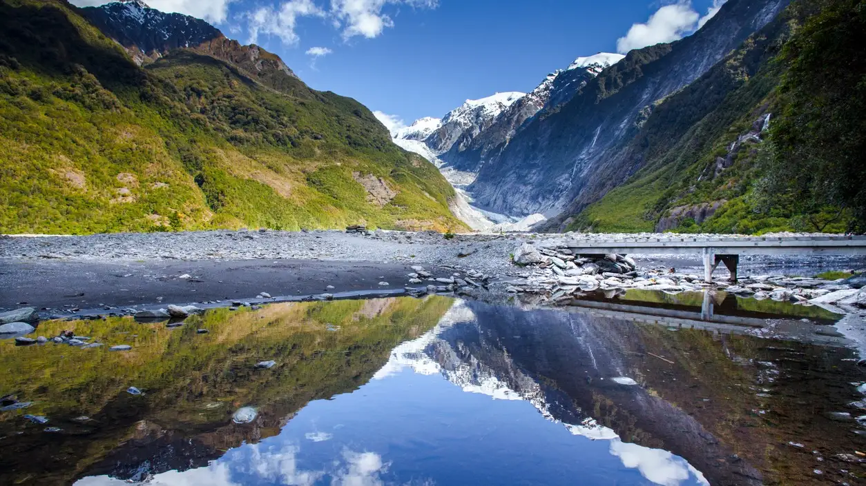 Der Franz-Joseph-Gletscher liegt zwischen zwei Bergen, die mit Gras bedeckt sind und im Vordergrund eine im Gewässer sichtbare Reflexion aufweisen.