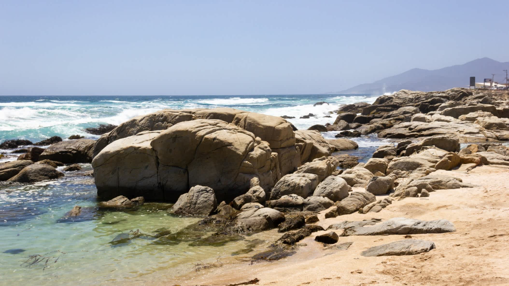 Der Strand Playa Aguas Blancas nahe Maitencillo in Chile bei sonnigem Wetter und mit schroffen Felsen im Vordergrund. 