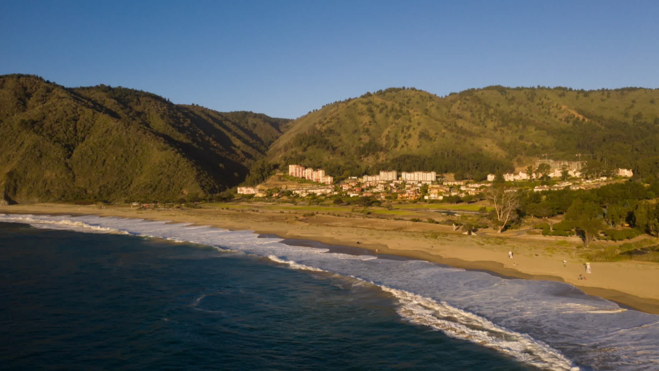 Der Luftblick auf den Strand Playa Grande in Chile bei blauem Himmel, mit Wellen im Wasser und dem Gebirge im Hintergrund sowie dem Ort Tongoy im Bild.