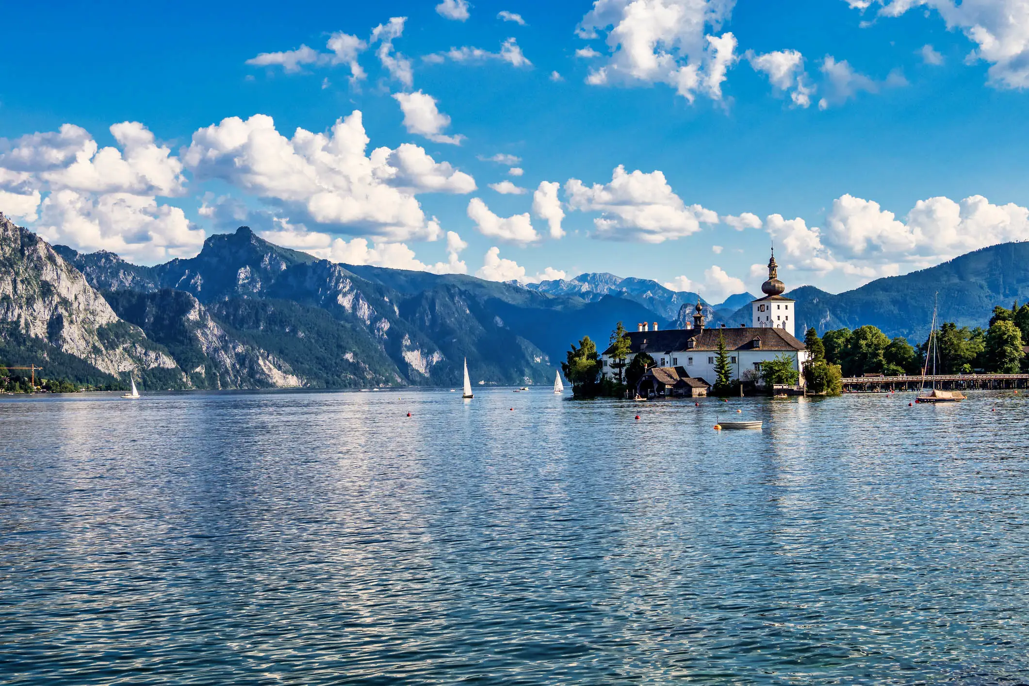 Scenic alpine lake with white castle on small island, sailboats on blue water, and mountains under fluffy clouds.