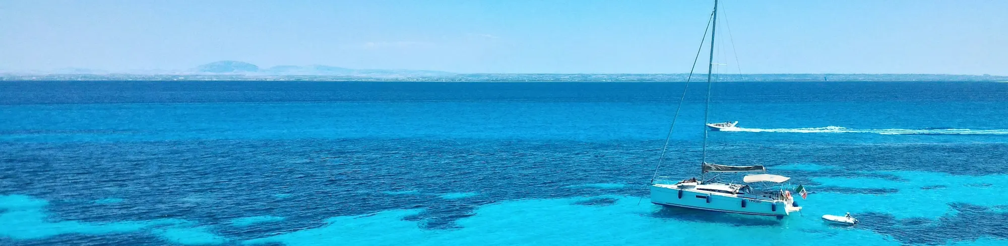 Türkises Meer mit strahlend blauem Himmel, im Vordergrund ist ein weißes Segelboot zu sehen, im Hintergrund zieht ein weißes Motorboot vorbei. 