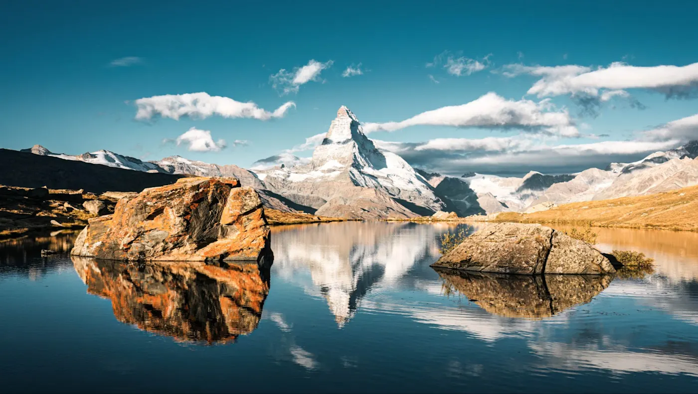The Matterhorn mountain reflected in a calm alpine lake with rocky outcrops under a bright blue sky with scattered clouds.
