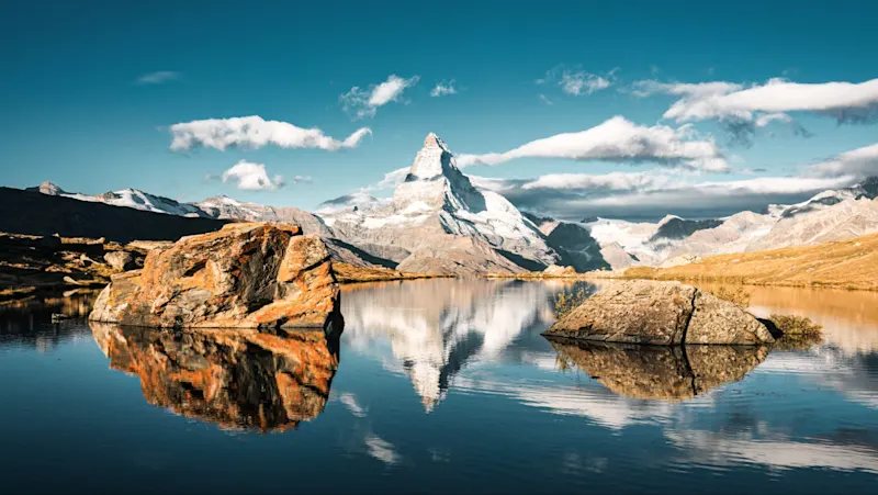 Zermatt, Switzerland The Matterhorn mountain reflected in a calm alpine lake with rocky outcrops under a bright blue sky with scattered clouds.
