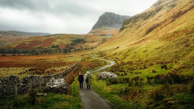Zwei Personen gehen einen gewundenen, von Steinmauern gesäumten Weg entlang, umgeben von grasbewachsenen Hügeln und Bergen unter einem bewölkten Himmel in einer malerischen ländlichen Landschaft.