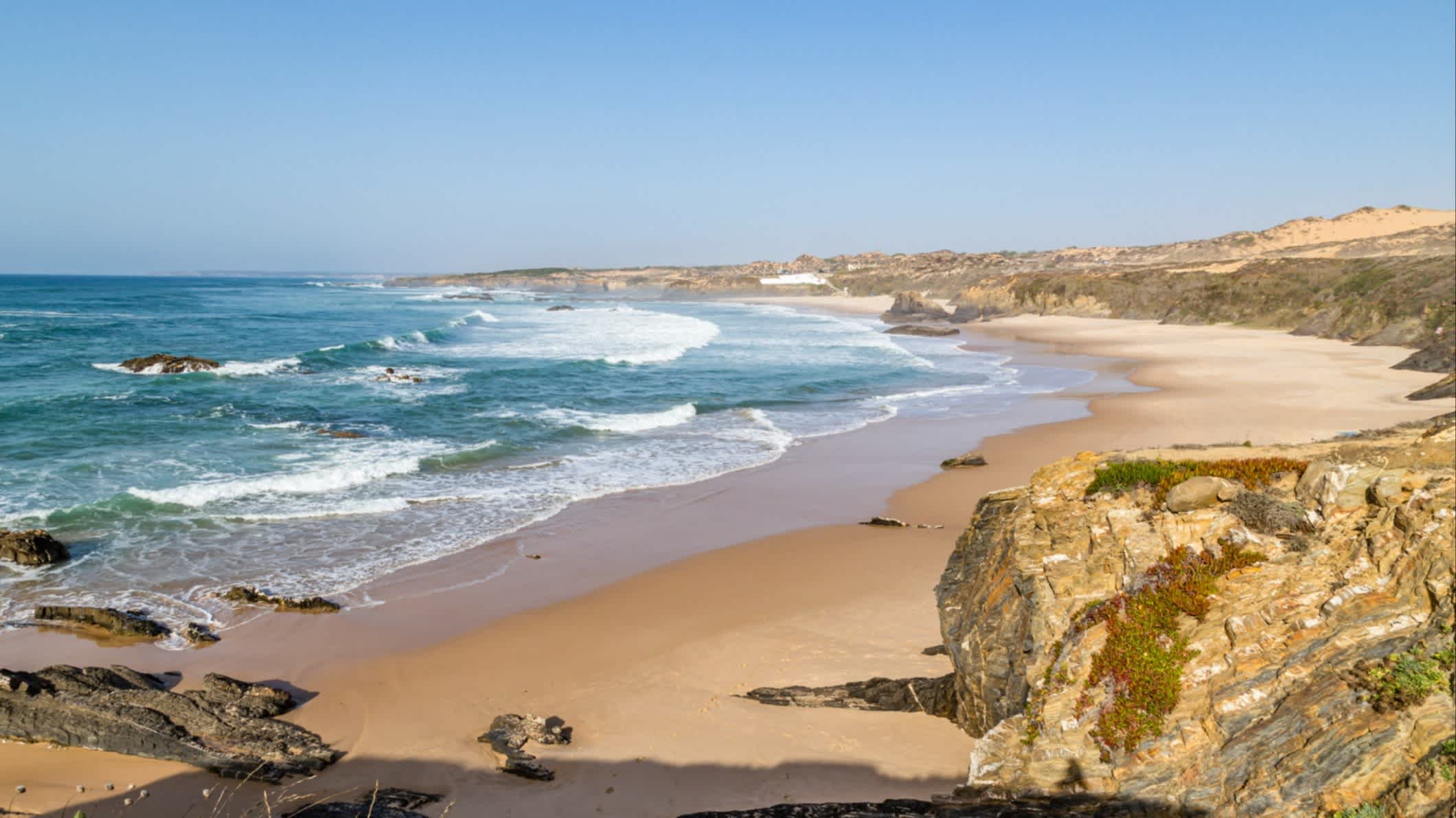 Der Strand Praia do Almograve, Alentejo, Portugal in der Bucht am Meer mit Felsen im Bild und bei herrlicher Sonne und Wellen im Wasser.