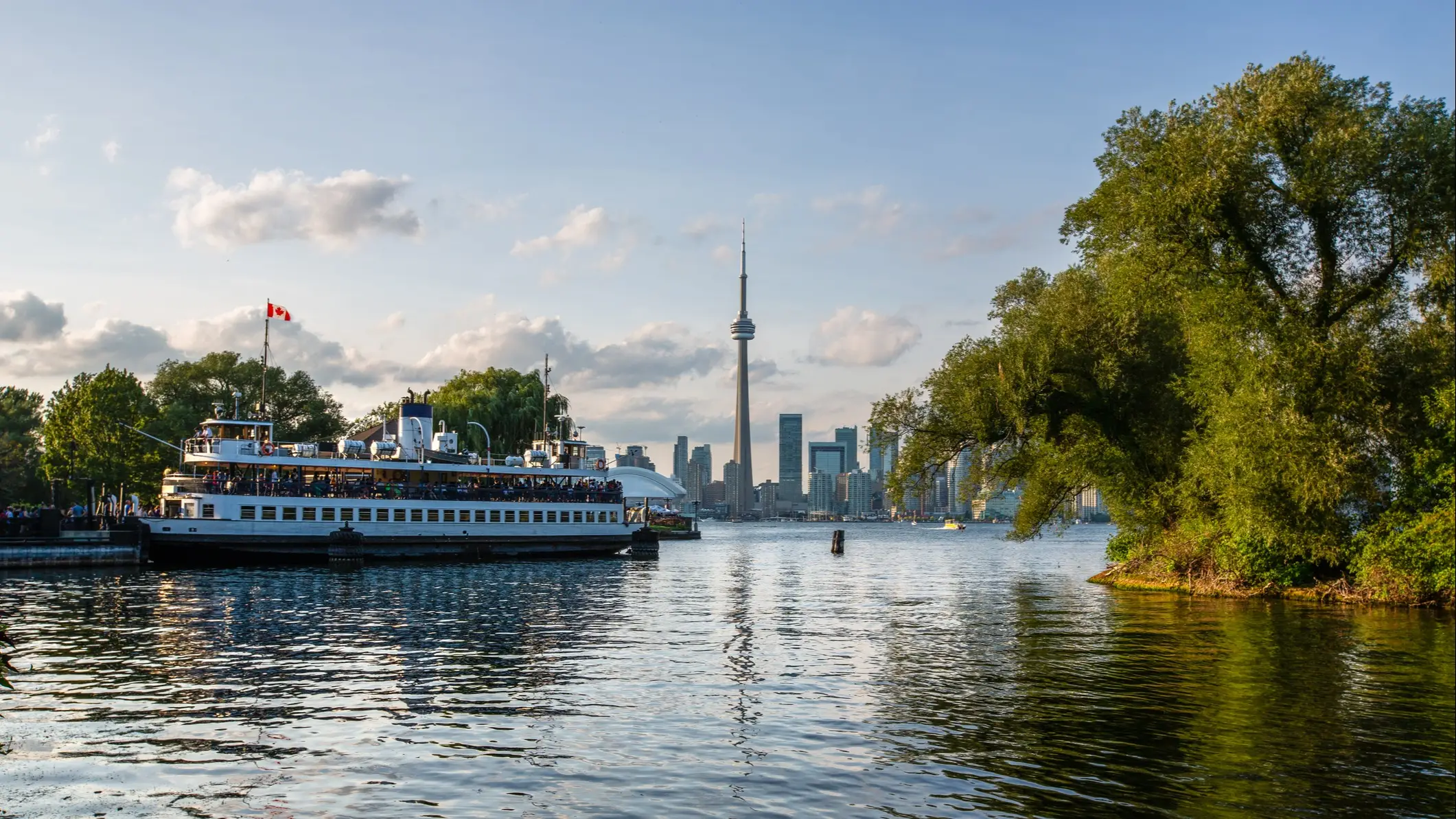 Ferry with Canadian flag docked near Toronto Islands with CN Tower and skyline in background under blue sky.