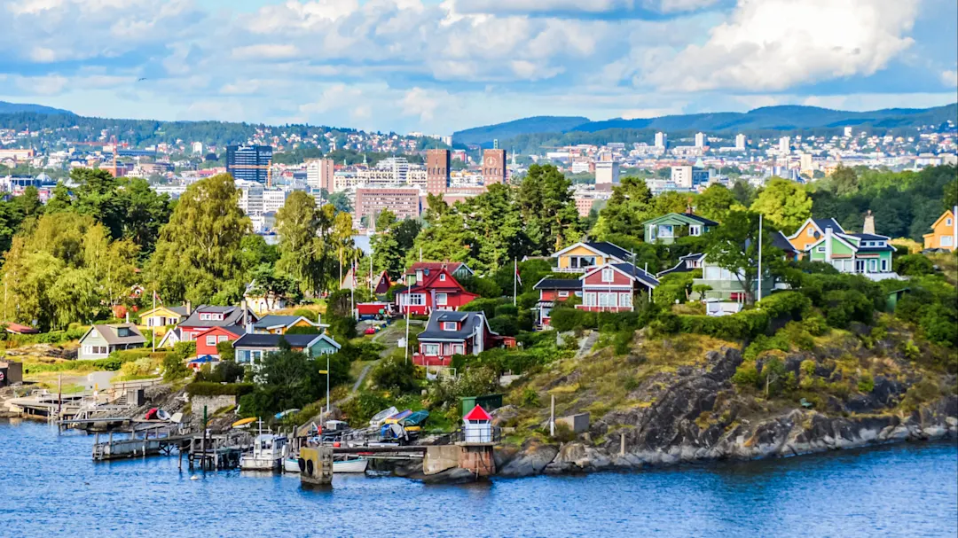 Panoramablick auf Oslo mit bunten Küstenhäusern am Wasser, Stadtgebäude im Hintergrund und bewaldeten Hügeln unter blauem Himmel.