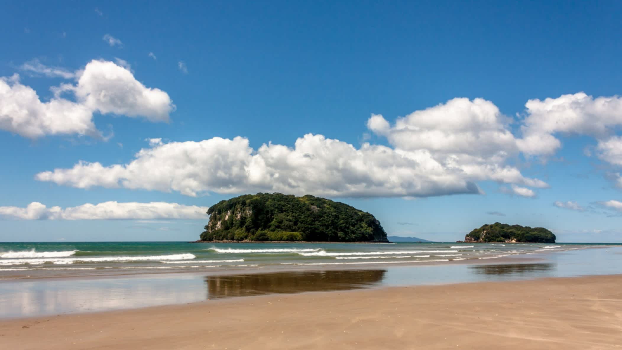 View of the sand and waves of Whangamata Beach, North Island, New Zealand with green rocks in the background

