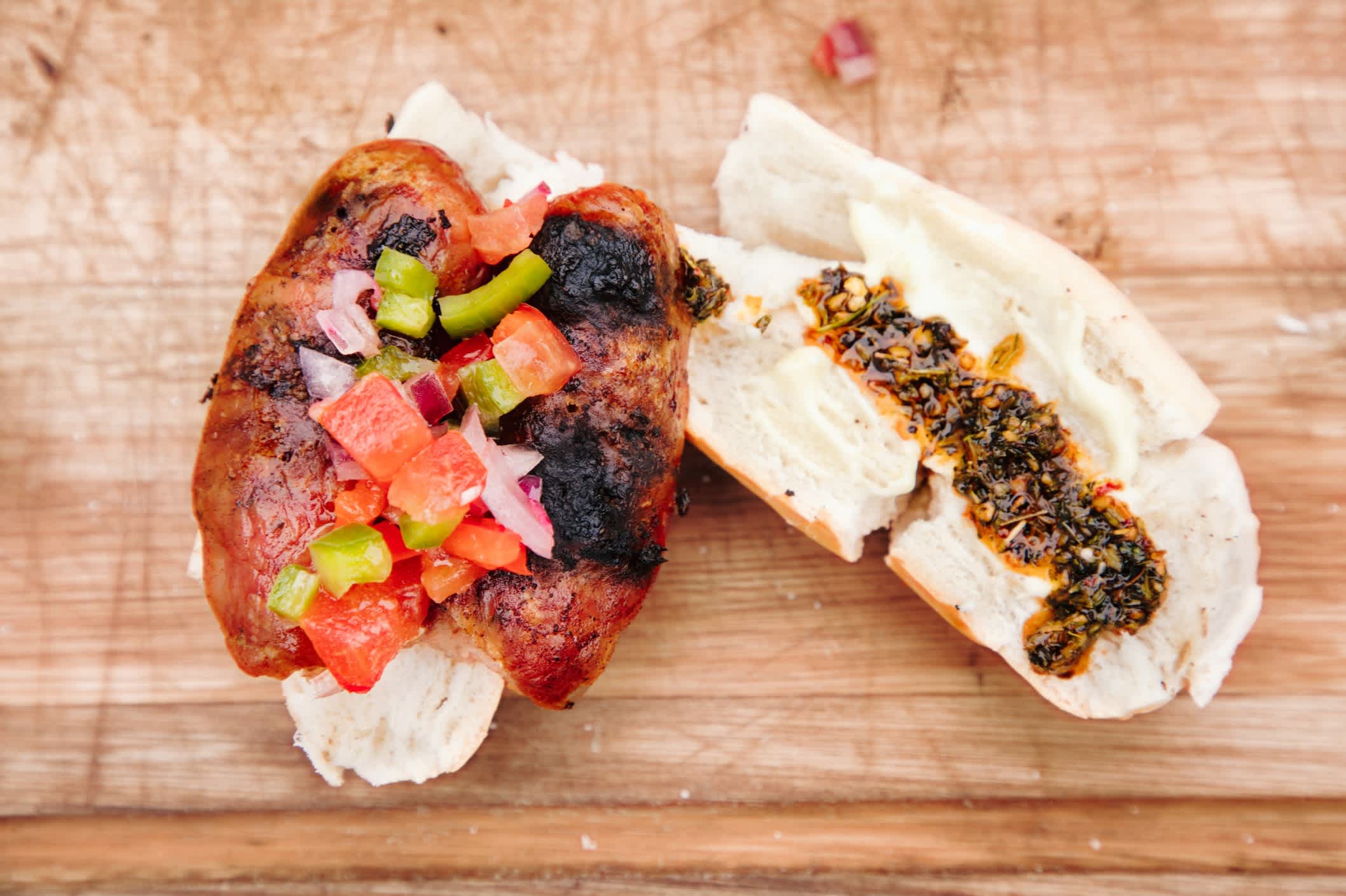 Von oben nach unten sehen Sie argentinisches Choripan Hot Dog Sandwich auf einem Schneidebrett, gewürzt mit Tomaten-Zwiebel-Salsa, Mayonnaise und Chimichurri.
