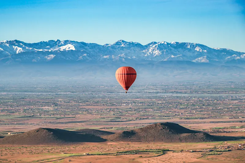 Ein roter Heißluftballon, der an einem klaren Frühlingstag über der Wüste in der Nähe von Marrakesch, Marokko, schwebt, mit dem schneebedeckten Atlasgebirge im Hintergrund.