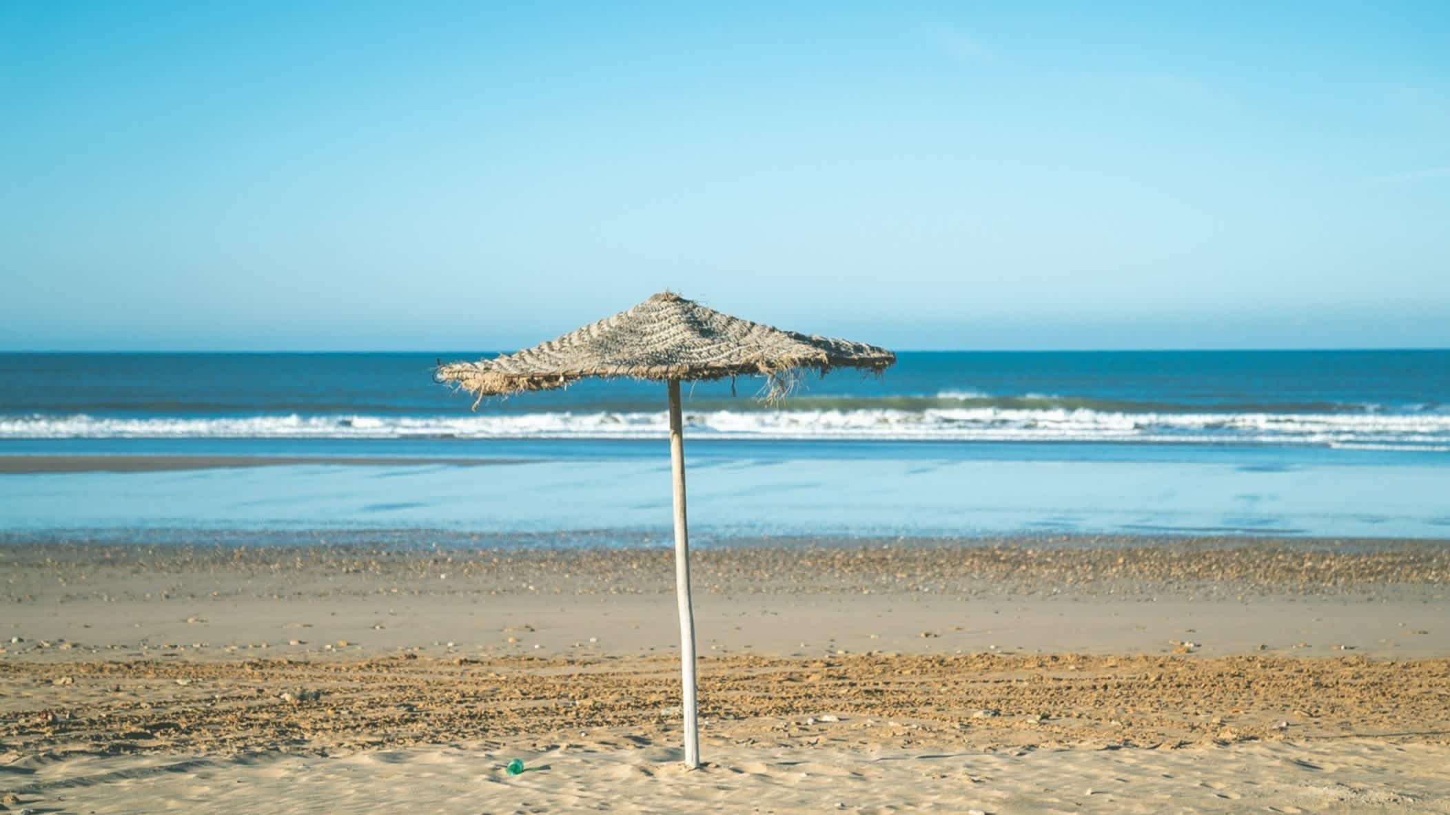 Ein Strandschirm am Strand von Sidi Bouzid, Marokko bei Sonnenschein und mit dem blauen Meer im Hintergrund.

