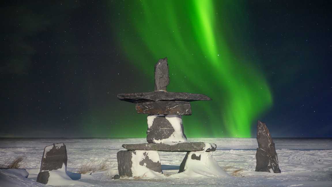 Un inukshuk en pierre en forme de croix dans la neige nocturne avec des aurores boréales vertes scintillantes en arrière-plan à Churchill, Canada.
