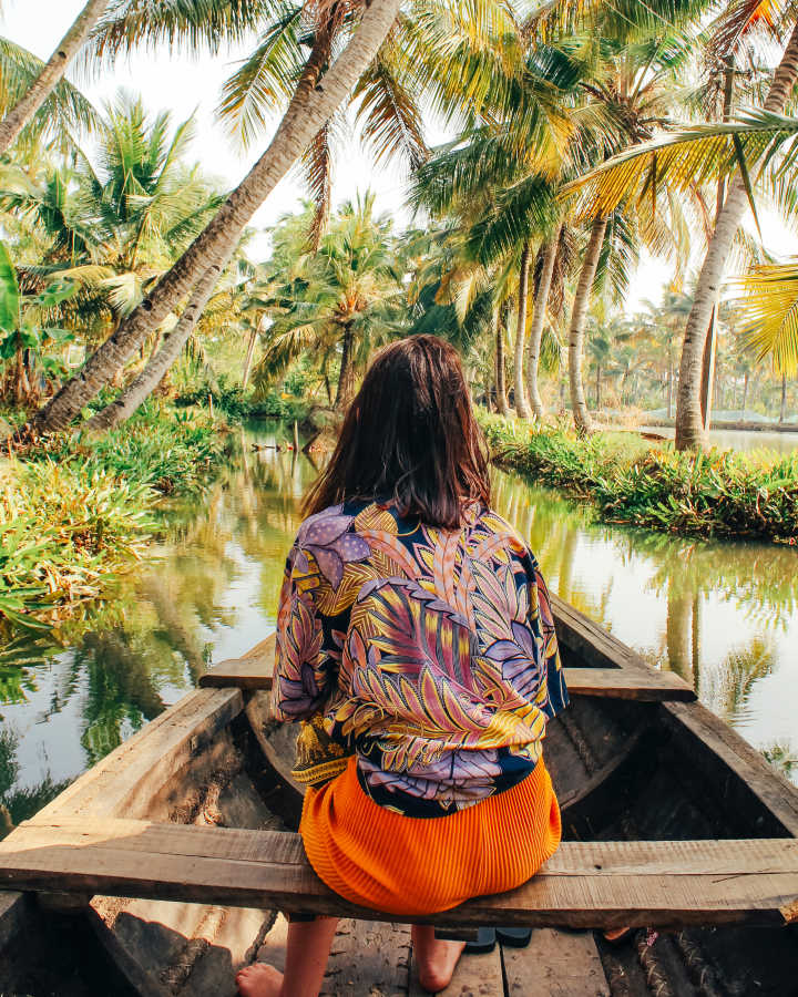 A woman from behind driving on a wooden boat through a river in the jungle.