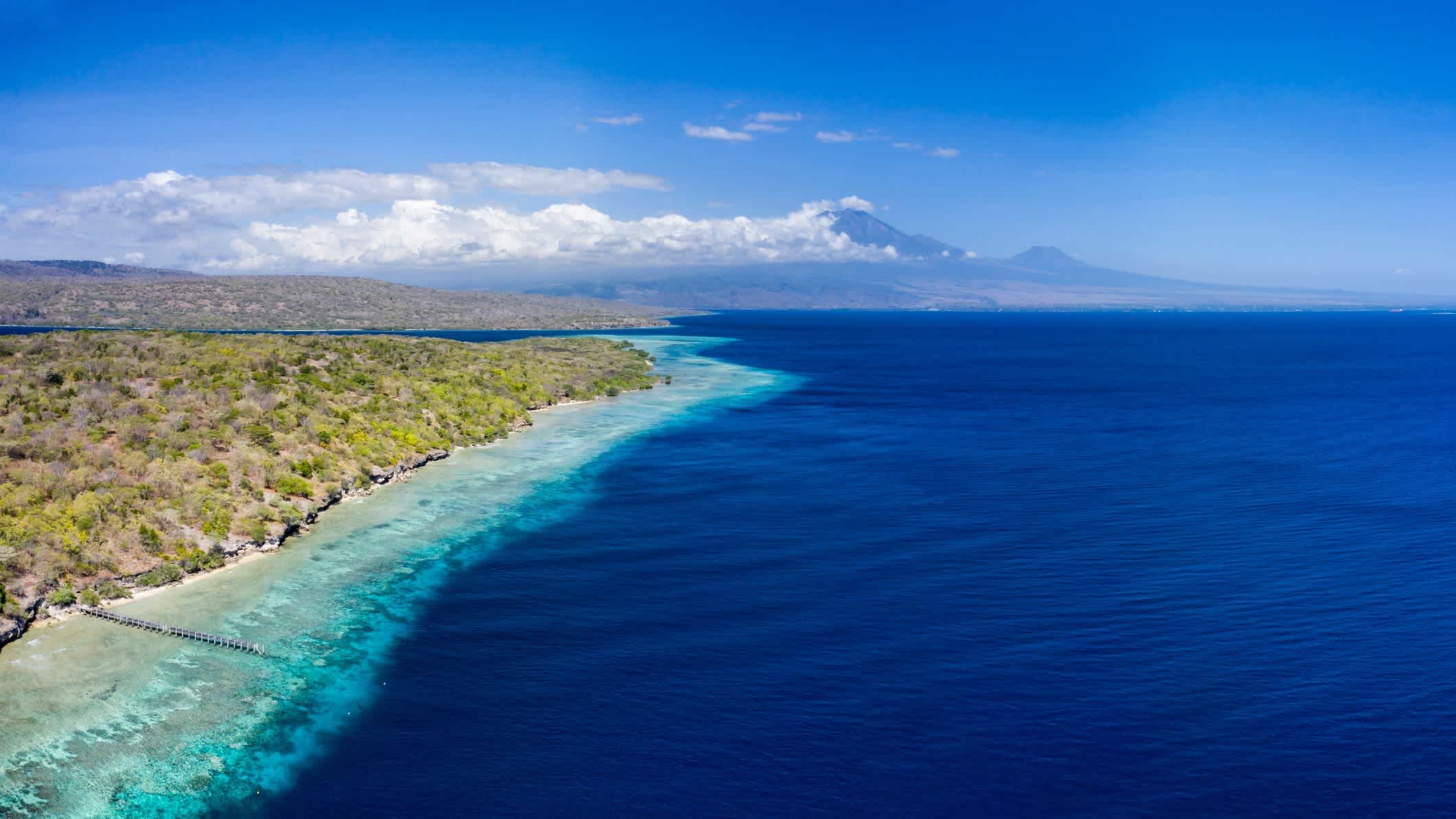 Luftaufnahme des tiefblauen Meers am weiten Sandstrand von Menjangan, Bali, Indonesien mit Blick auf die Berge im Hintergrund.