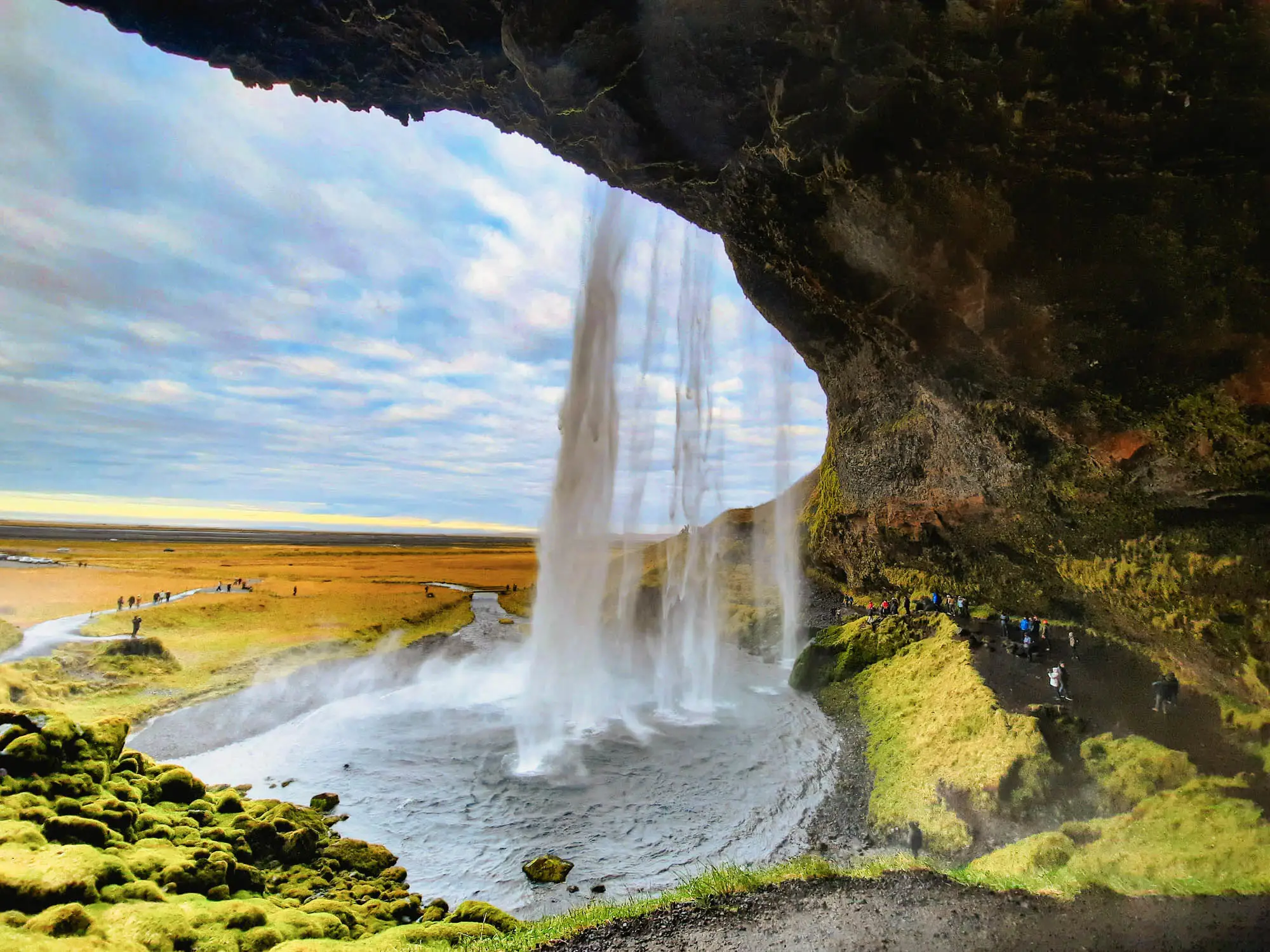 Blick hinter einen Wasserfall in Island mit weiter Landschaft und blauem Himmel, Besucher erkunden den Pfad dahinter.
