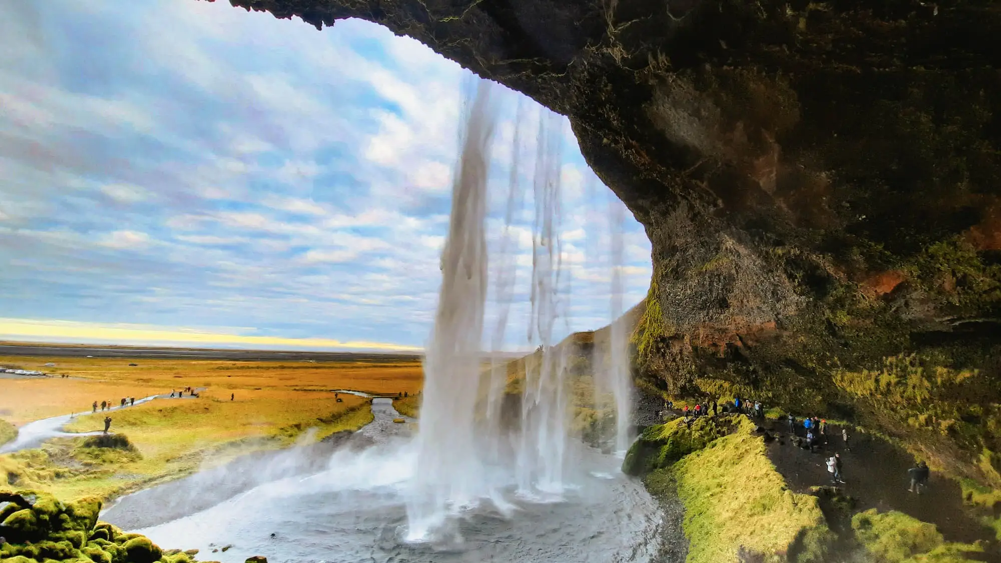 Blick hinter einen Wasserfall in Island mit weiter Landschaft und blauem Himmel, Besucher erkunden den Pfad dahinter.