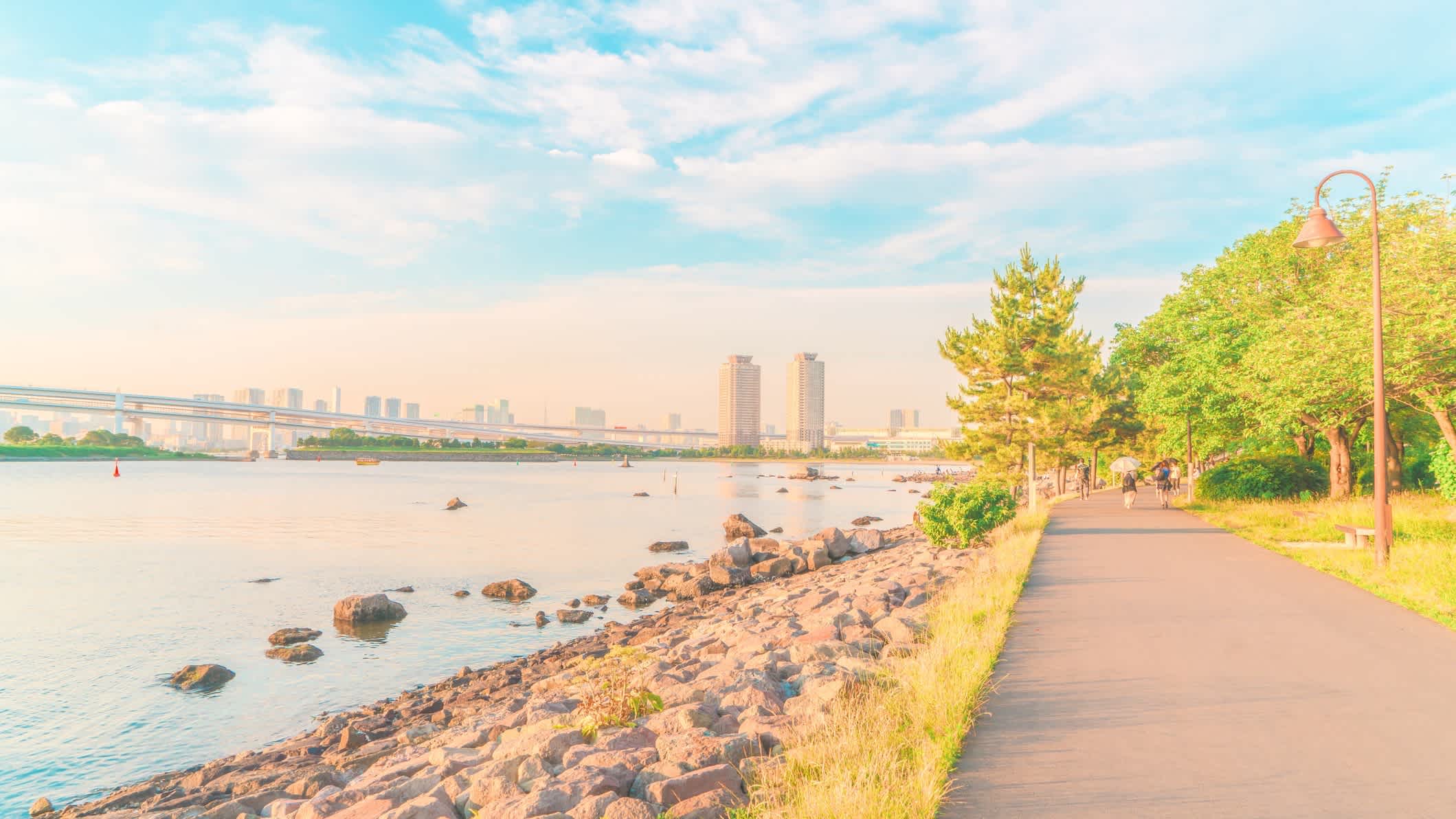 Spazierweg am Wasser von Odaiba, Tokyo Bay, Tokio, Japan bei sonnigem Wetter und mit Blick auf die Stadt sowie den Stadtpark.