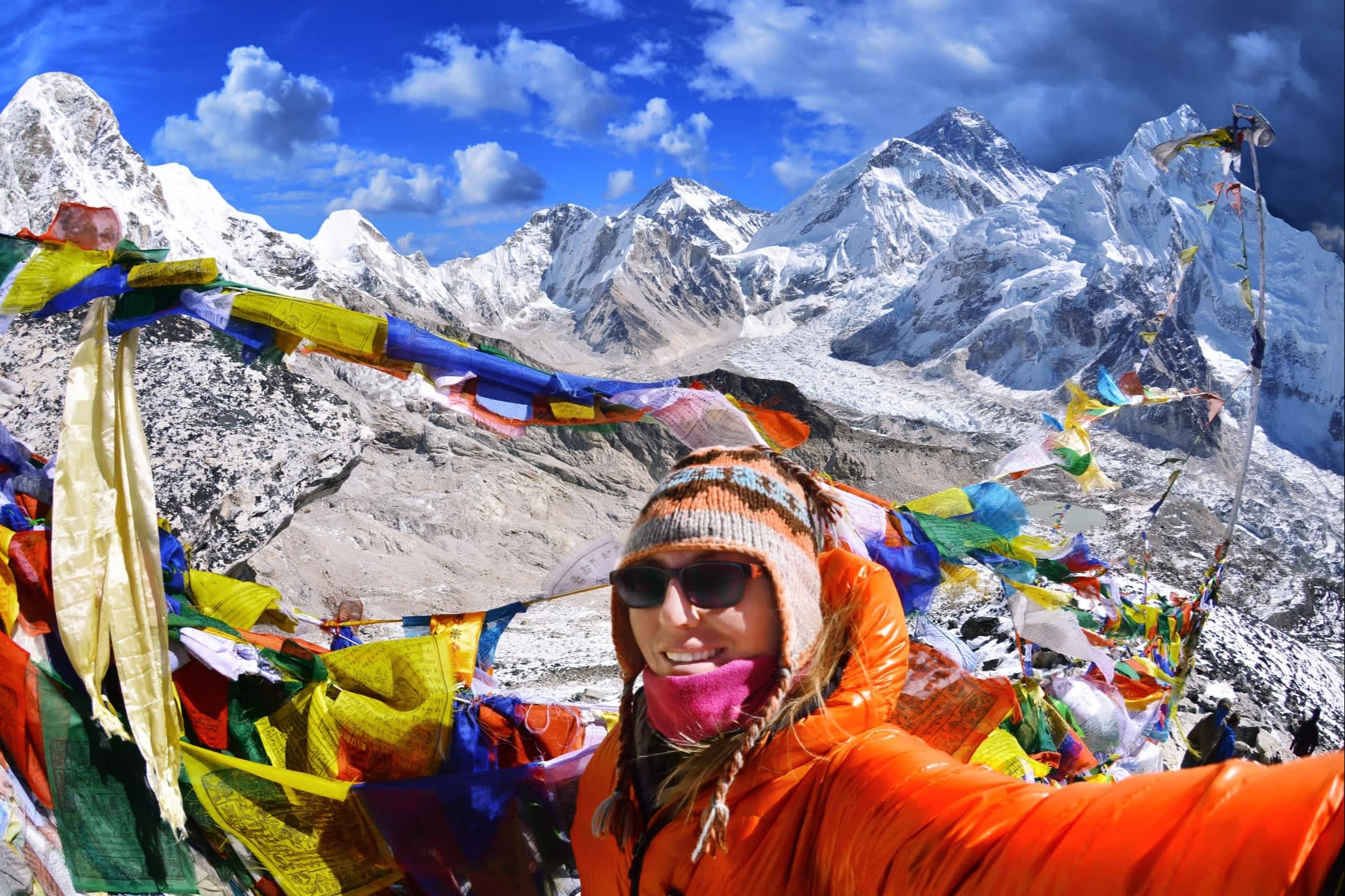 Mount Everest Camp in Nepal mit einer Frau, die ein Selfie macht an einem sonnigen Tag mit den typischen nepalesischen Fahnen im Hintergrund und verschneite Gipfel des Himalayas.