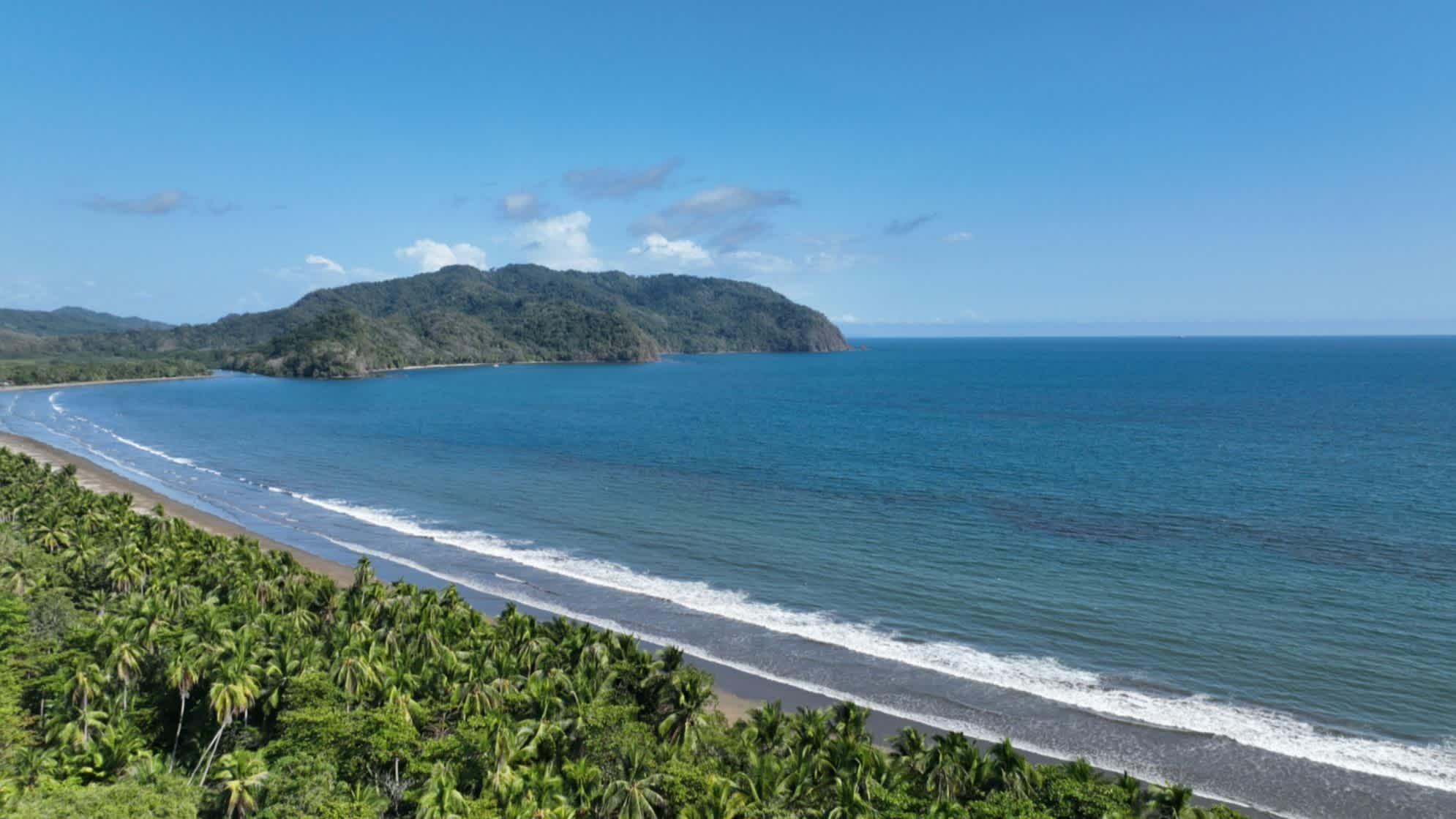 Der Strand Playa Tambor auf der Nicoya-Halbinsel in Costa Rica bei blauem Himmel und mit seichten Wellen im Wasser sowie grünen Hügeln im Hintergrund.