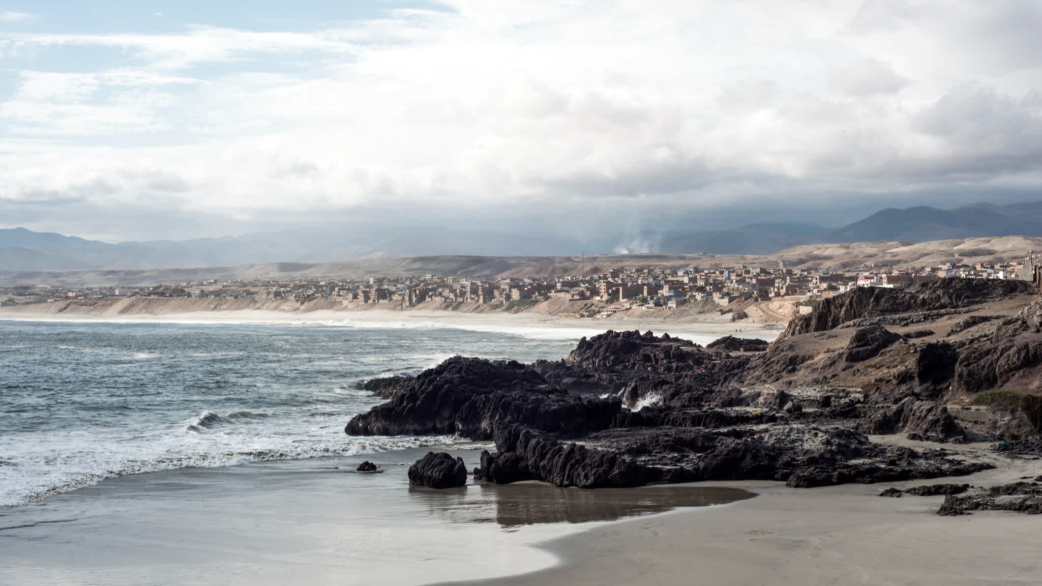 Ein natürlicher Strand in Chala, Arequipa, Peru bei Sonne-Wolken-Mix und mit einer Sand-Felsen-Küste sowie einem Gebirge im Hintergrund.